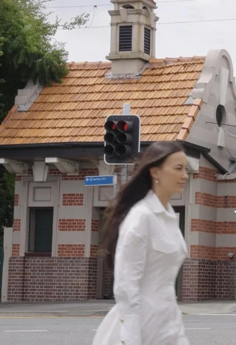 A woman with long dark hair, wearing a white coat, walking across a street near a traffic light and a small brick building with a tiled roof.