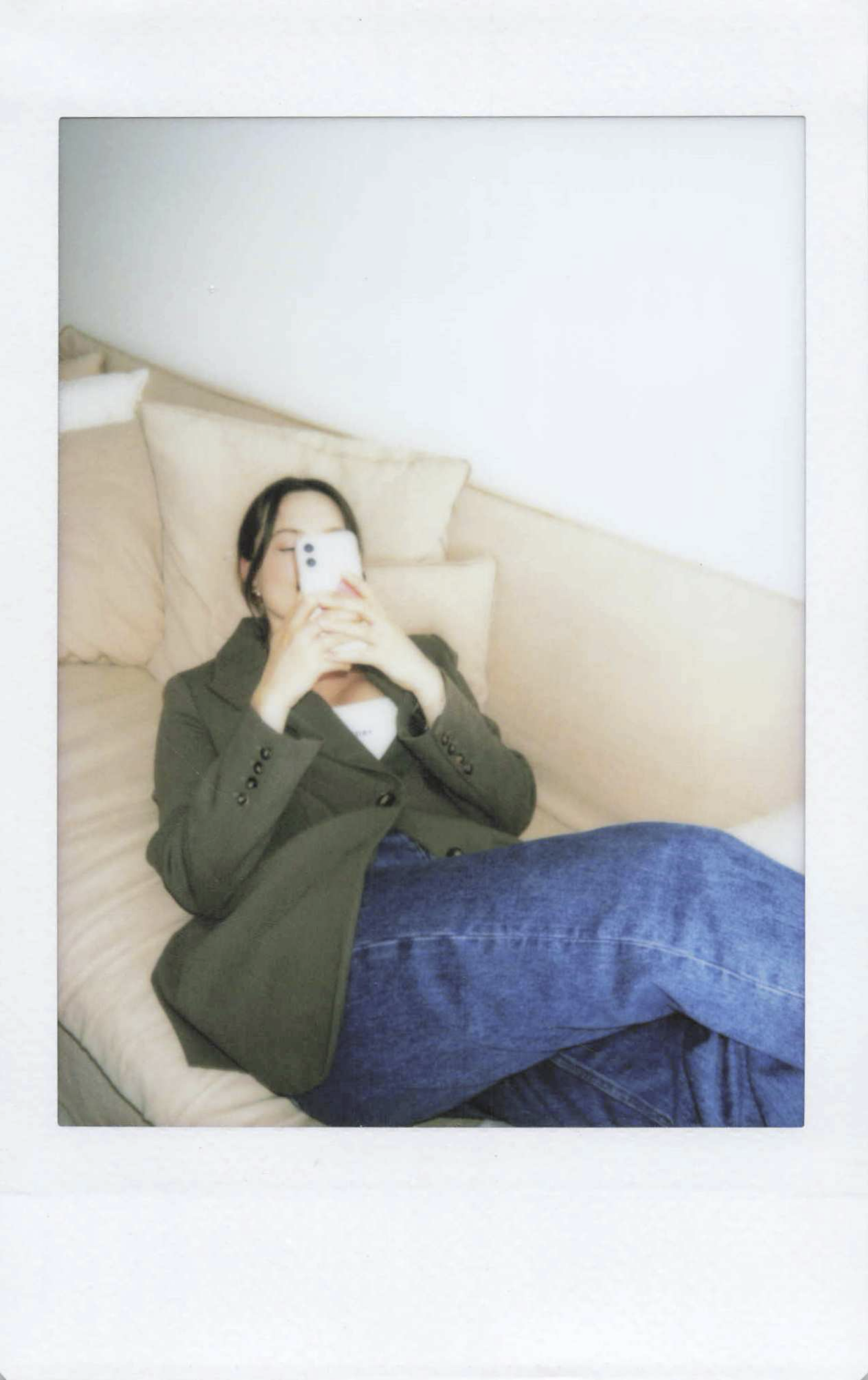 A woman is lying on a beige couch on her smartphone, wearing a green jacket and blue jeans.