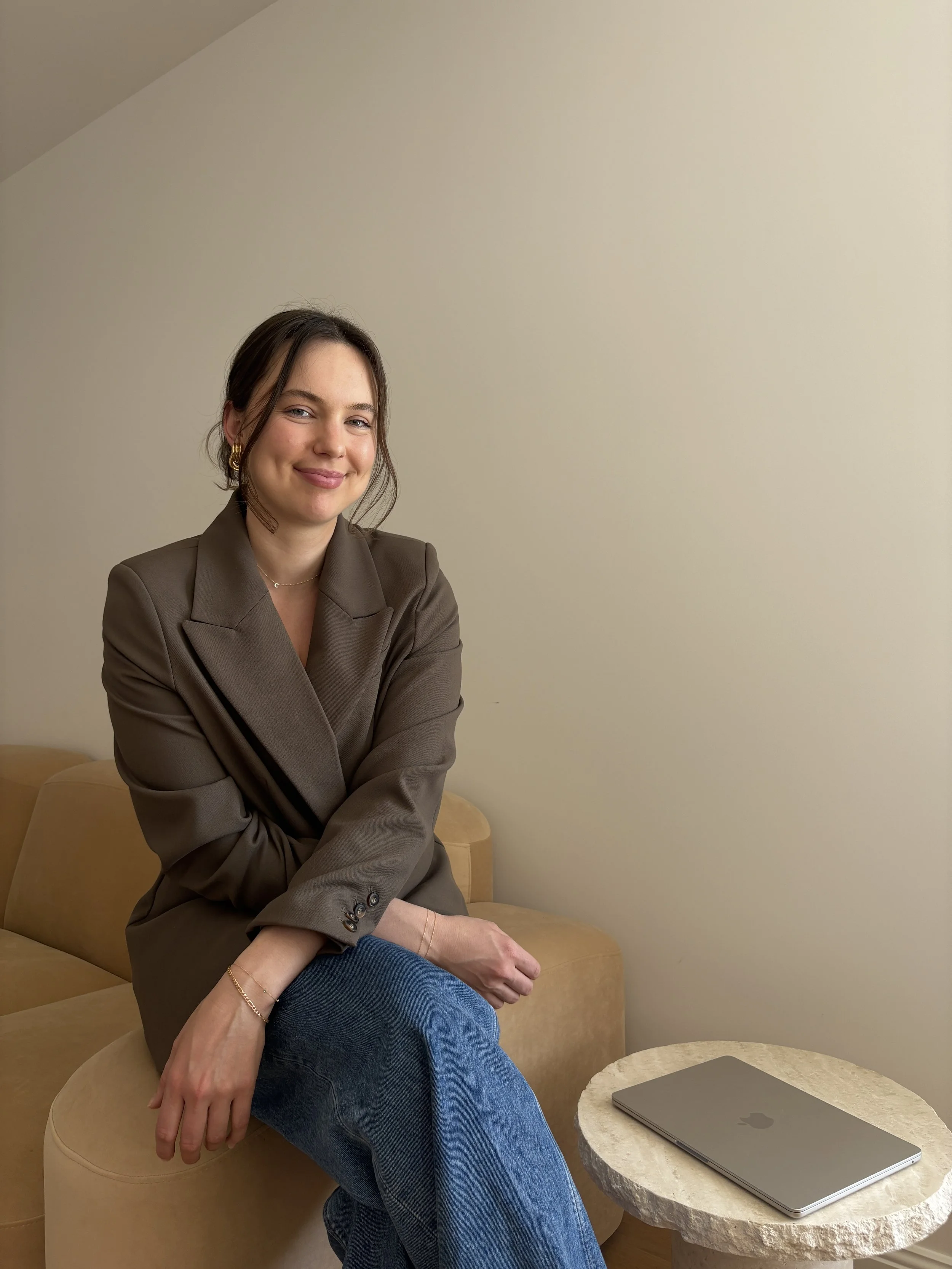 A young woman sitting on a beige sofa with a closed MacBook laptop on a small round table next to her, in a room with plain light-colored walls, smiling at the camera.