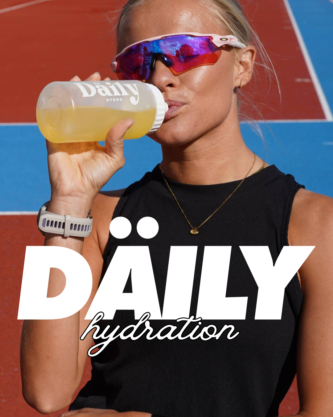 A woman wearing sunglasses and a black sleeveless top drinks from a large water bottle labeled "Daily Hydro" after exercise on a red and blue athletic track.