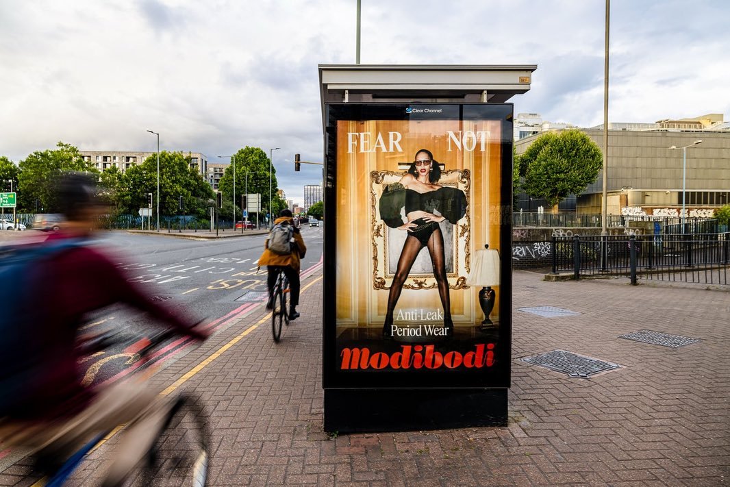 An outdoor advertisement on a bus stop featuring a woman in tights and black clothing with the text 'FEAR NOT' and 'Modibodi Anti-Leak Period Wear.'