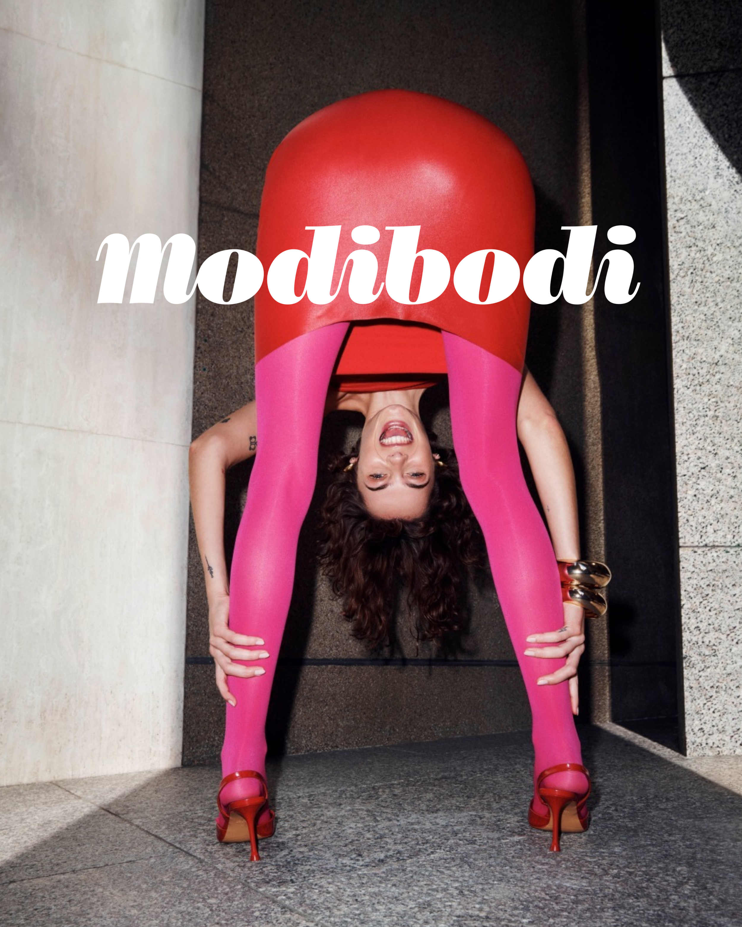A woman with curly hair in a pose with her head between her legs, wearing pink tights and red high heels, with a red skirt,, and large bracelets on her wrists, against a tiled wall background.