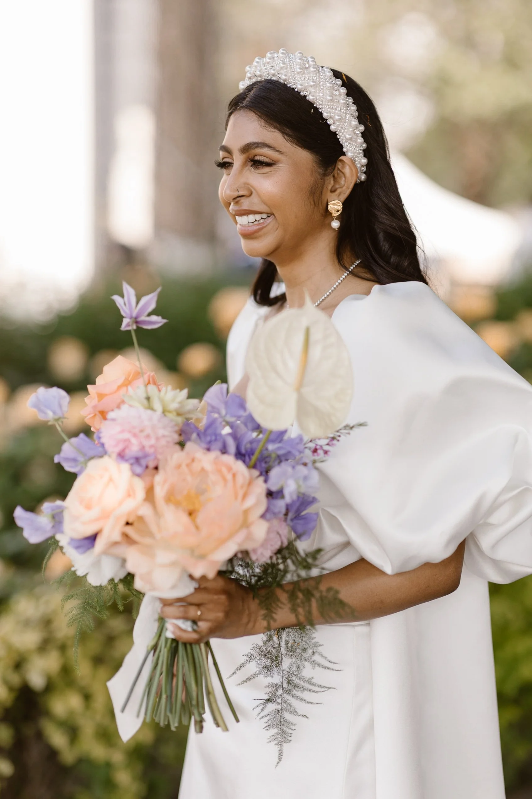 A bride smiling, holding a colorful bouquet of flowers, wearing a white gown, pearl and gold earrings, and a pearl headband, outdoors with blurred trees and tents in the background.