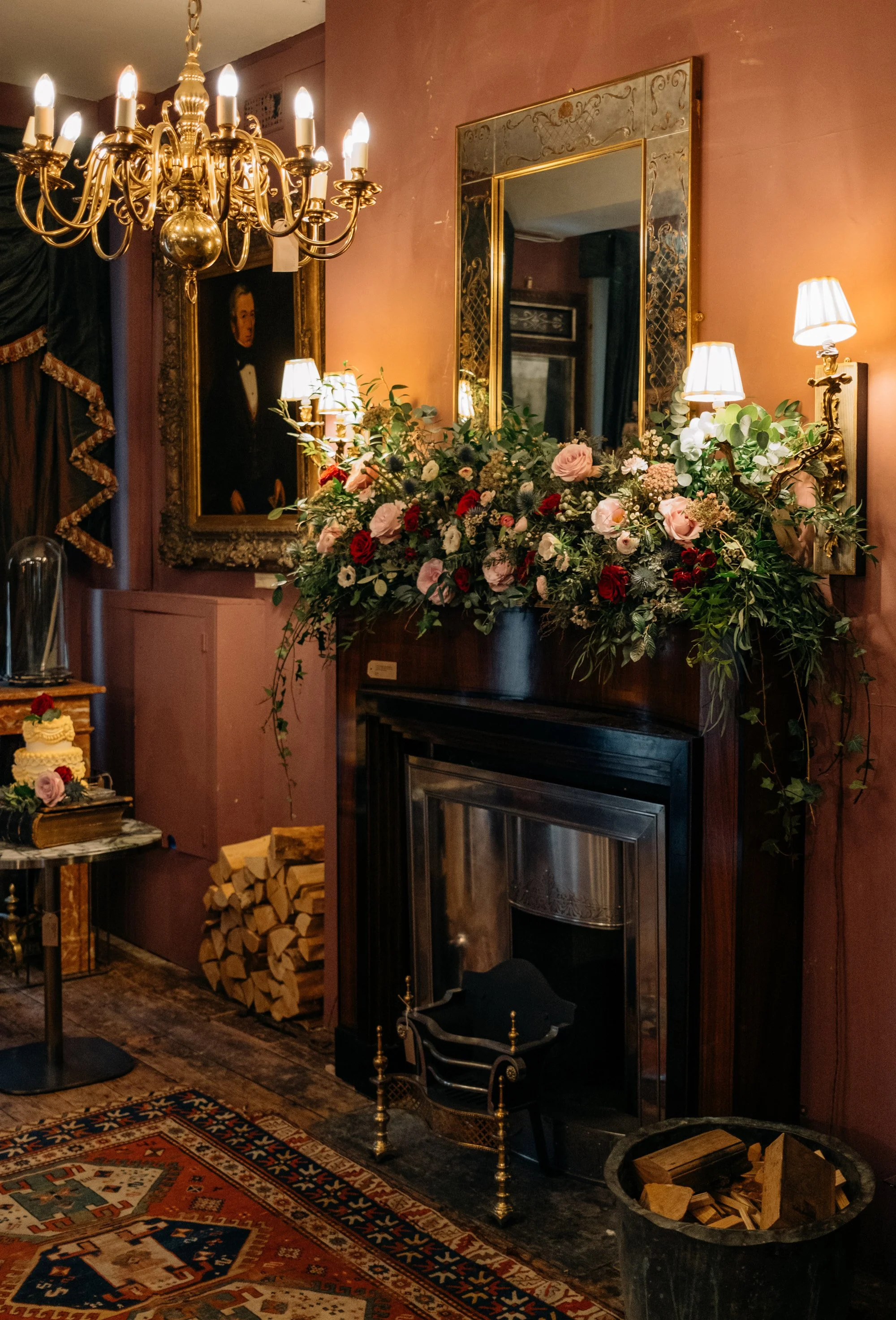 An elegant living room corner with a fireplace decorated with a large floral arrangement, a mirror, wall sconces, a chandelier, a portrait painting, and a patterned rug.