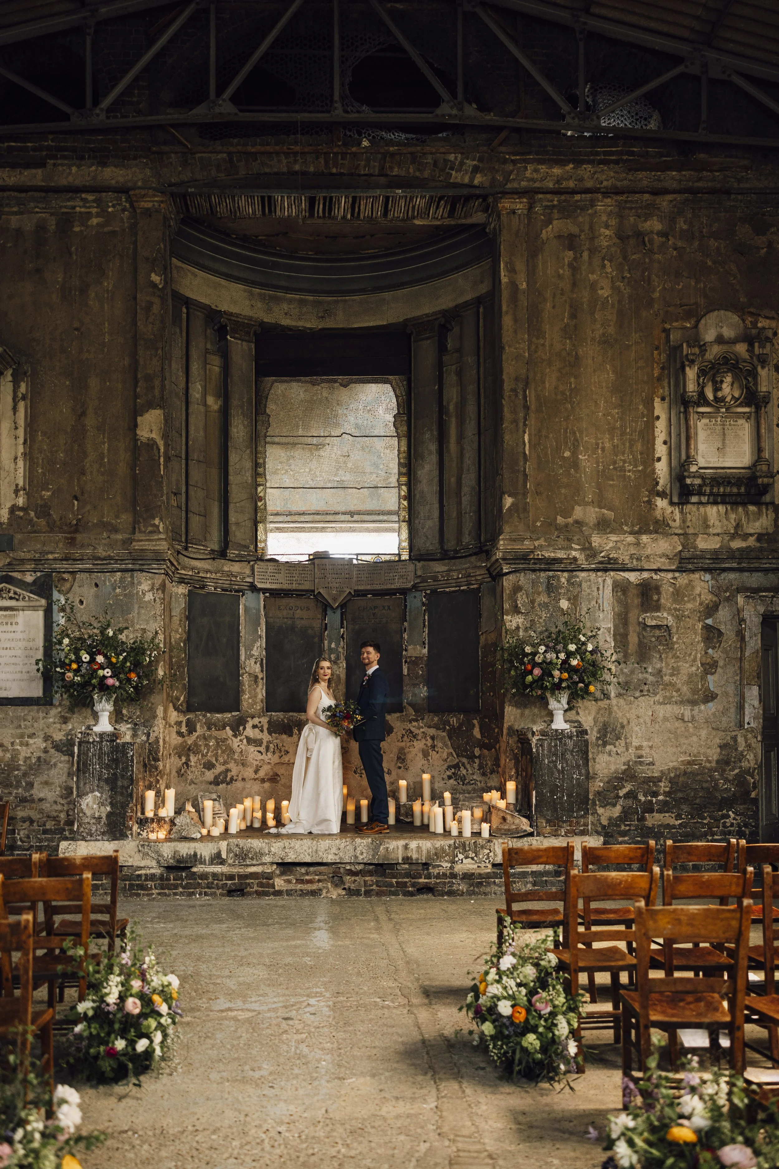 A bride and groom standing on a stone platform in a rustic, historic church with candles and flower arrangements, during their wedding.