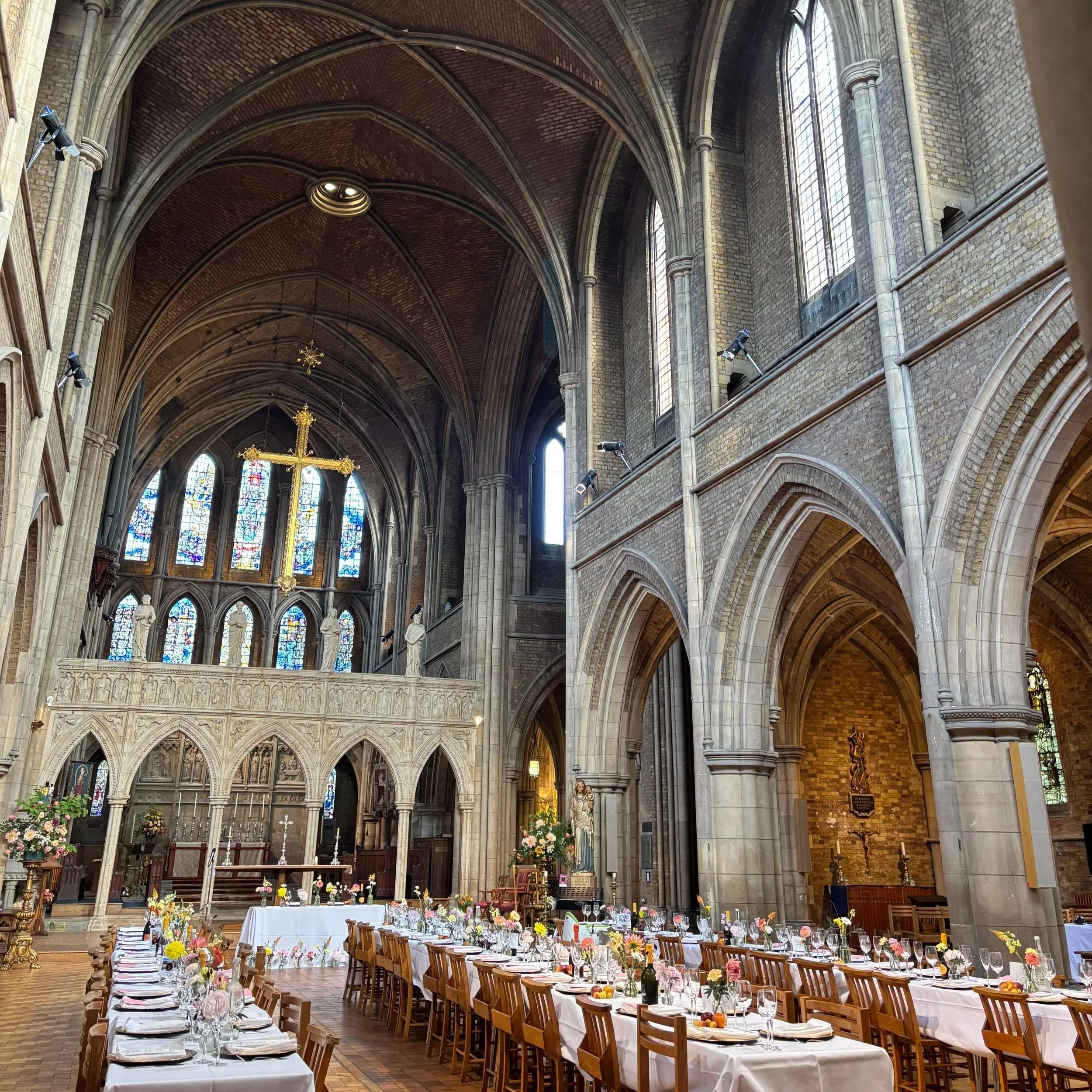 Interior of a large church decorated for a celebration with long banquet tables covered in white tablecloths, floral centerpieces, and set with plates, glasses, and cutlery, with stained glass windows and high vaulted ceilings.