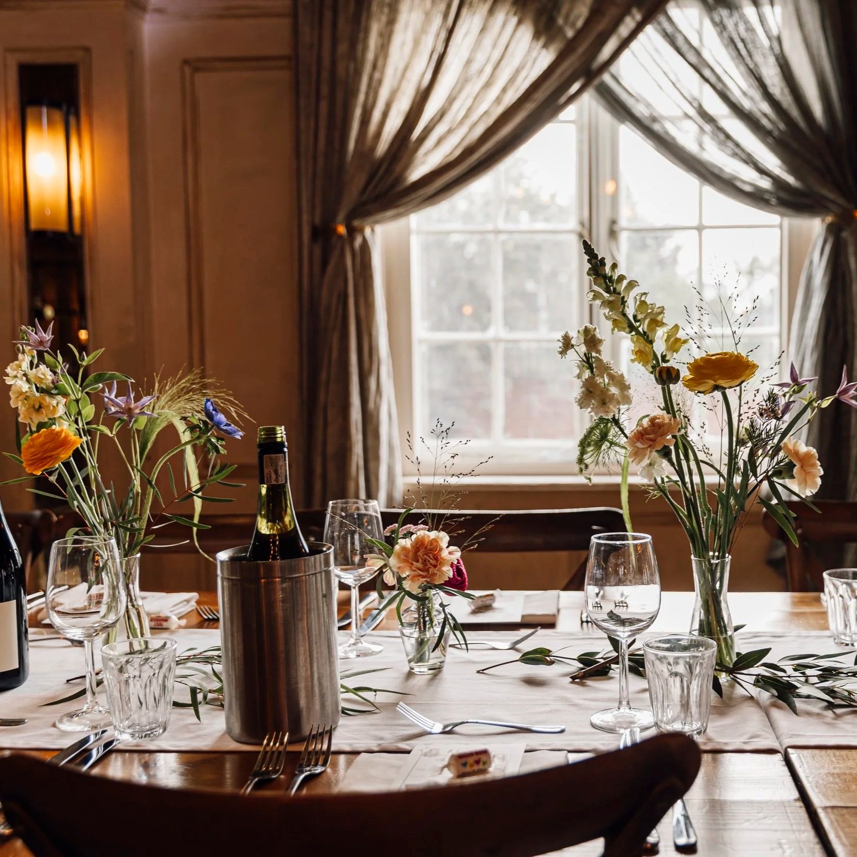 A dining table set with floral centerpieces, glassware, and a wine bottle in a cozy room with wooden paneling, curtains, and a large window letting in natural light.