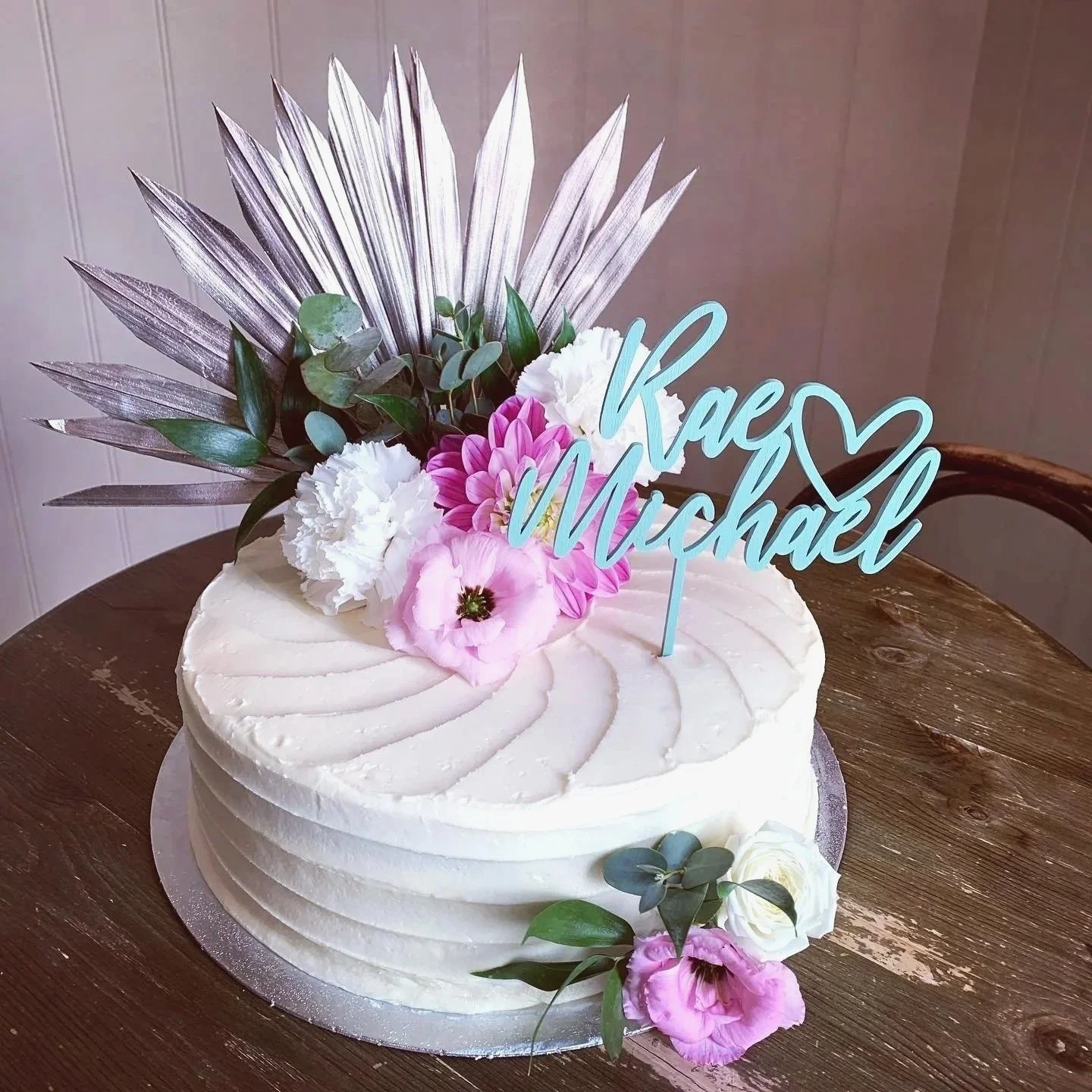 White frosted birthday cake decorated with pink and white flowers, green leaves, and silver leaves, with a blue 'Kaci y Michael' cake topper and a heart symbol, placed on a wooden table.