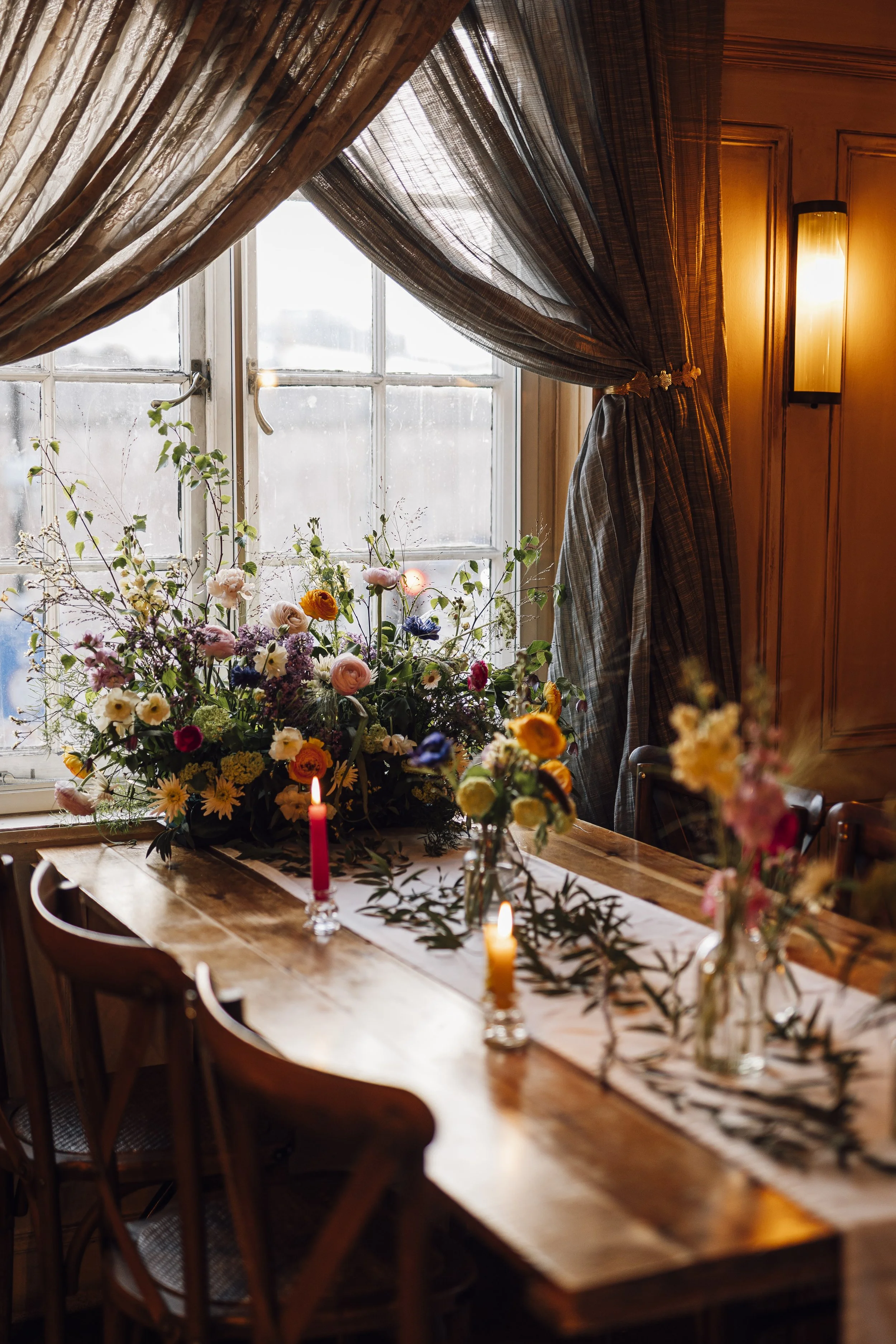 A wooden dining table decorated with vases of colorful flowers, lit candles, and a white table runner, set in front of a window with draped curtains and warm wall lighting.