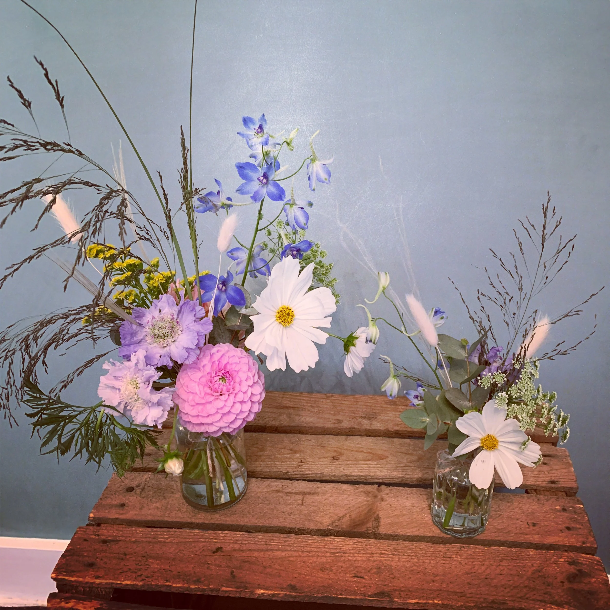 Two glass vases with colorful flowers and greenery on a wooden table, against a light blue wall.