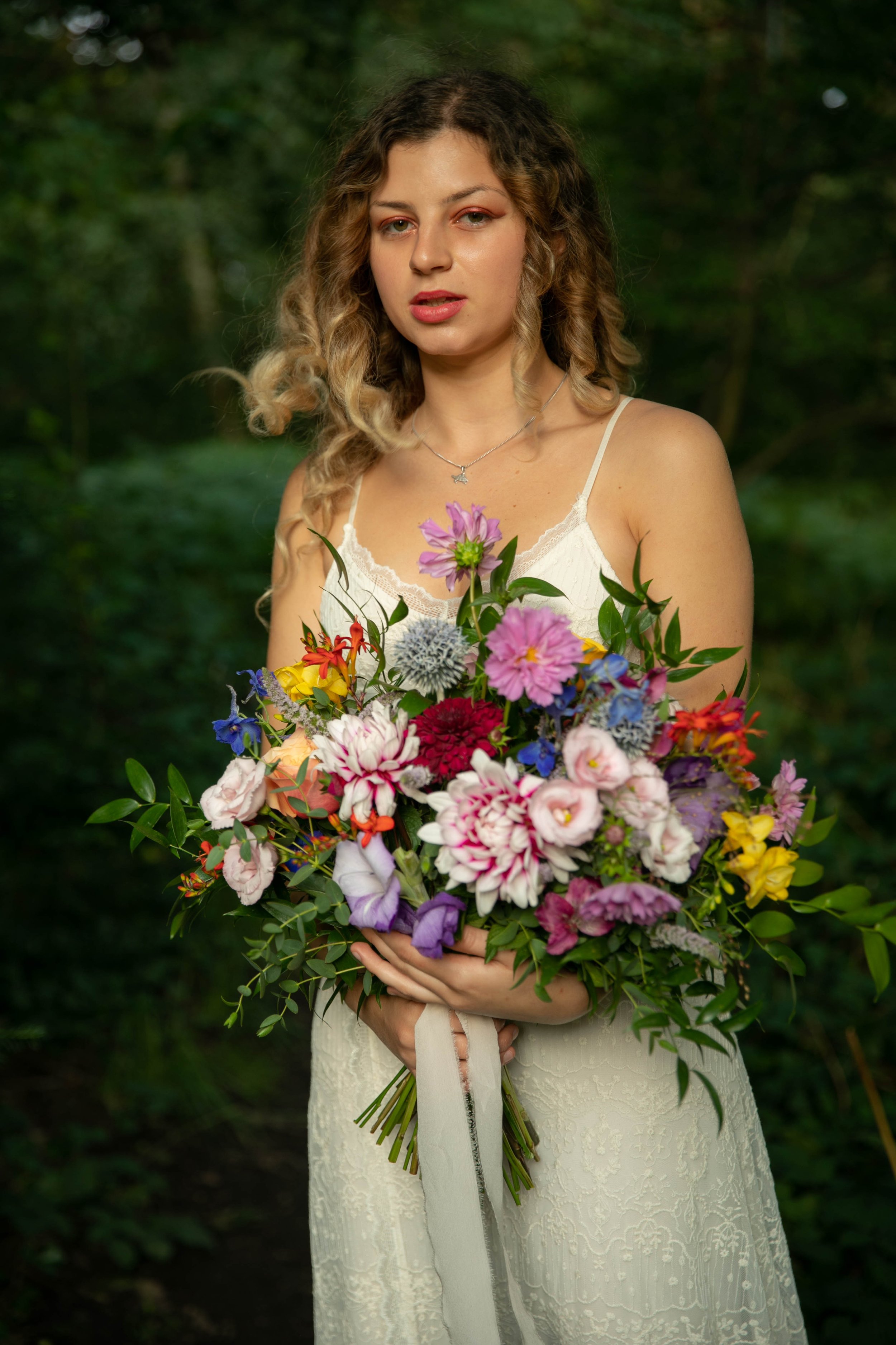 A woman with wavy blonde hair in a white lace dress holding a large colorful bouquet of flowers outdoors with a green, wooded background.