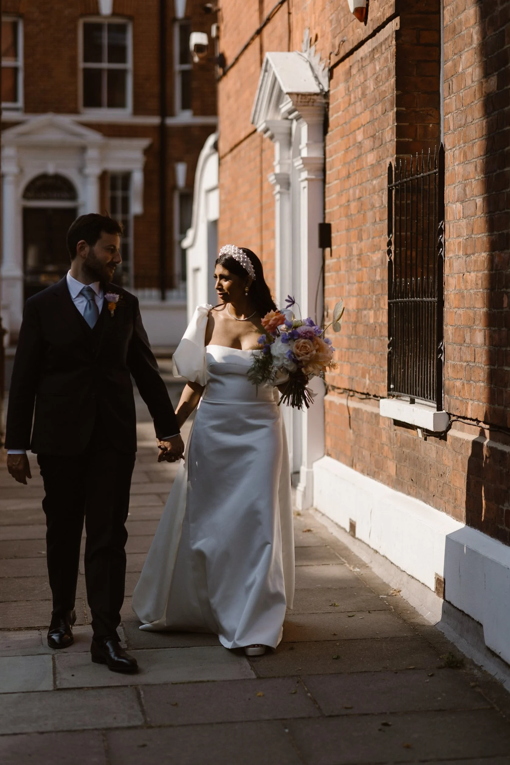 A bride and groom walking hand in hand on a city sidewalk, with the bride holding a bouquet of flowers, both dressed in wedding attire during daytime.