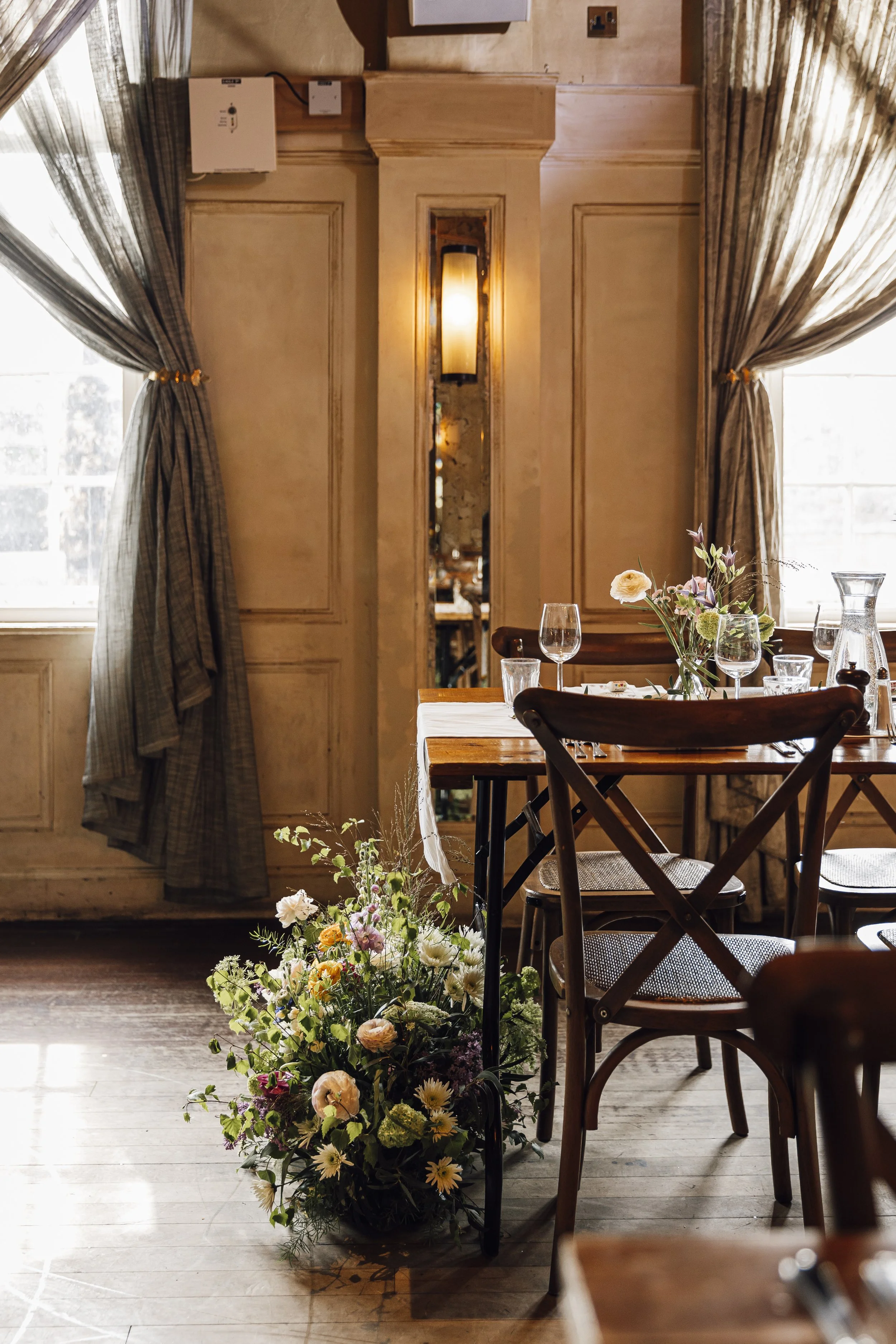 A dining room with natural light, featuring a table set with glasses and a floral centerpiece, and a large floral arrangement on the floor underneath.