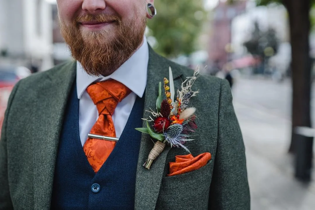 Close-up of a man with a beard wearing a gray suit with a boutonniere, an orange tie, and a pocket square, standing outdoors.