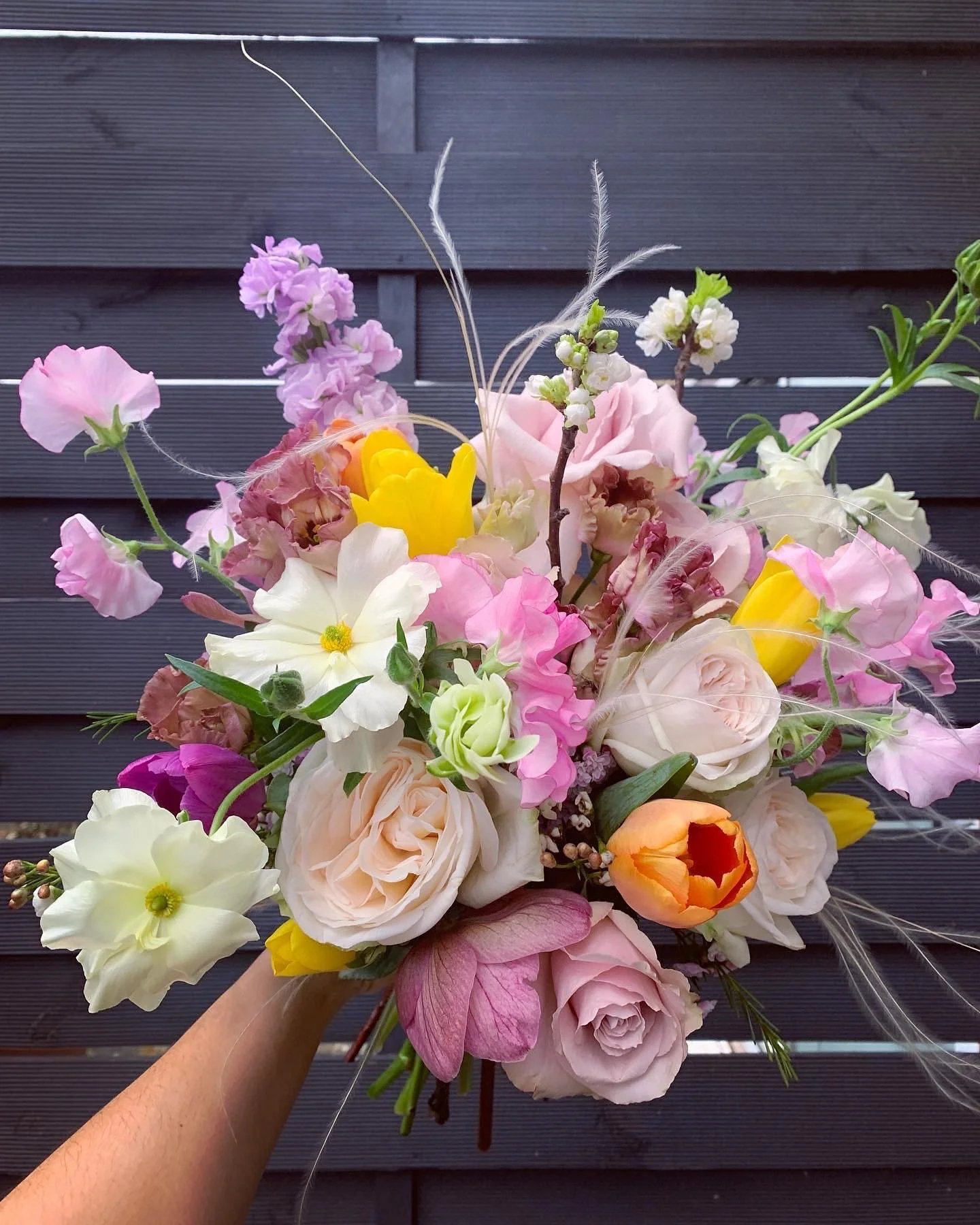 A hand holding a colorful bouquet of various flowers including roses, tulips, and other blooms, with a dark wooden background.