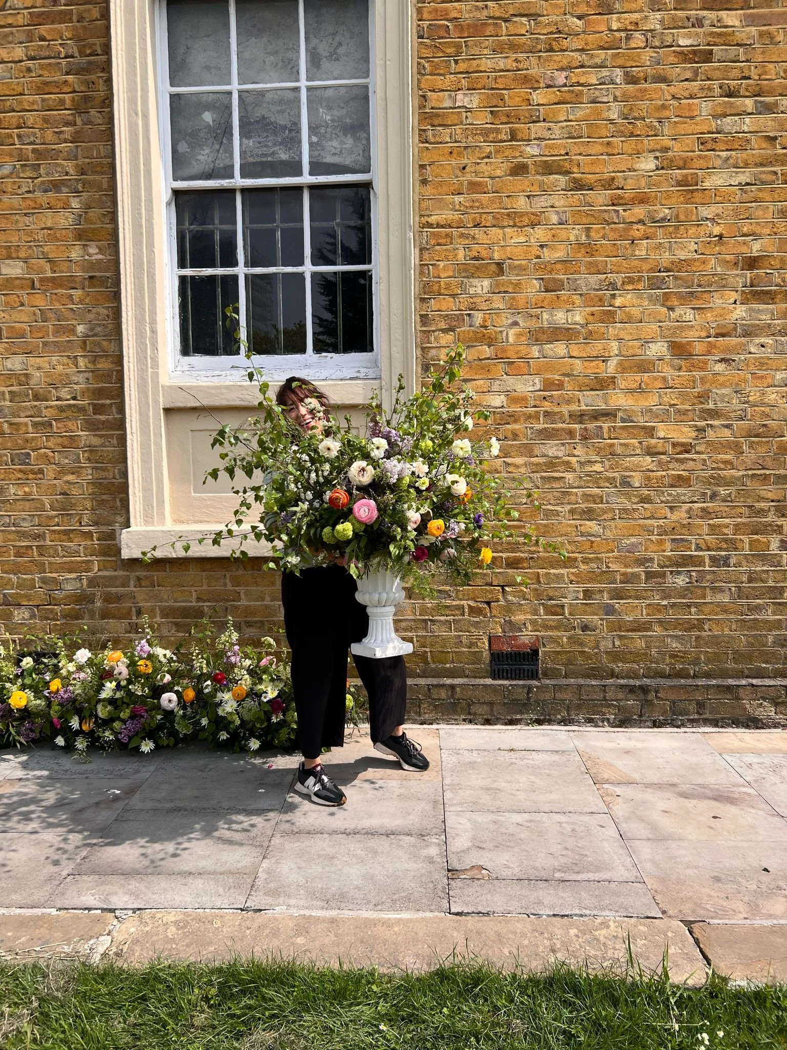 Person standing behind a large floral arrangement in a white vase, smiling, outdoors against a brick wall with a window.