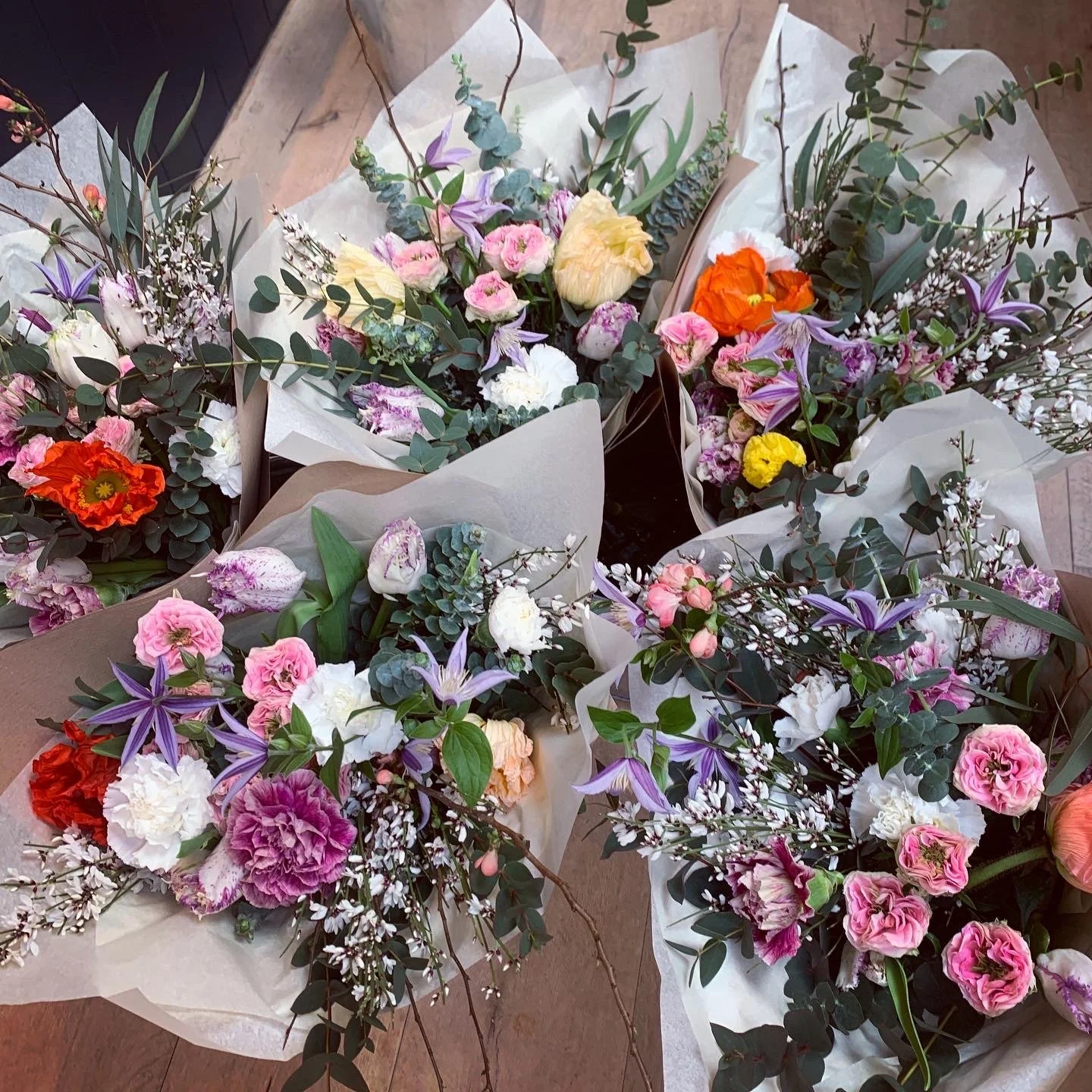 Multiple colorful flower bouquets with pink, purple, white, yellow, and orange flowers wrapped in paper, arranged on a wooden surface.