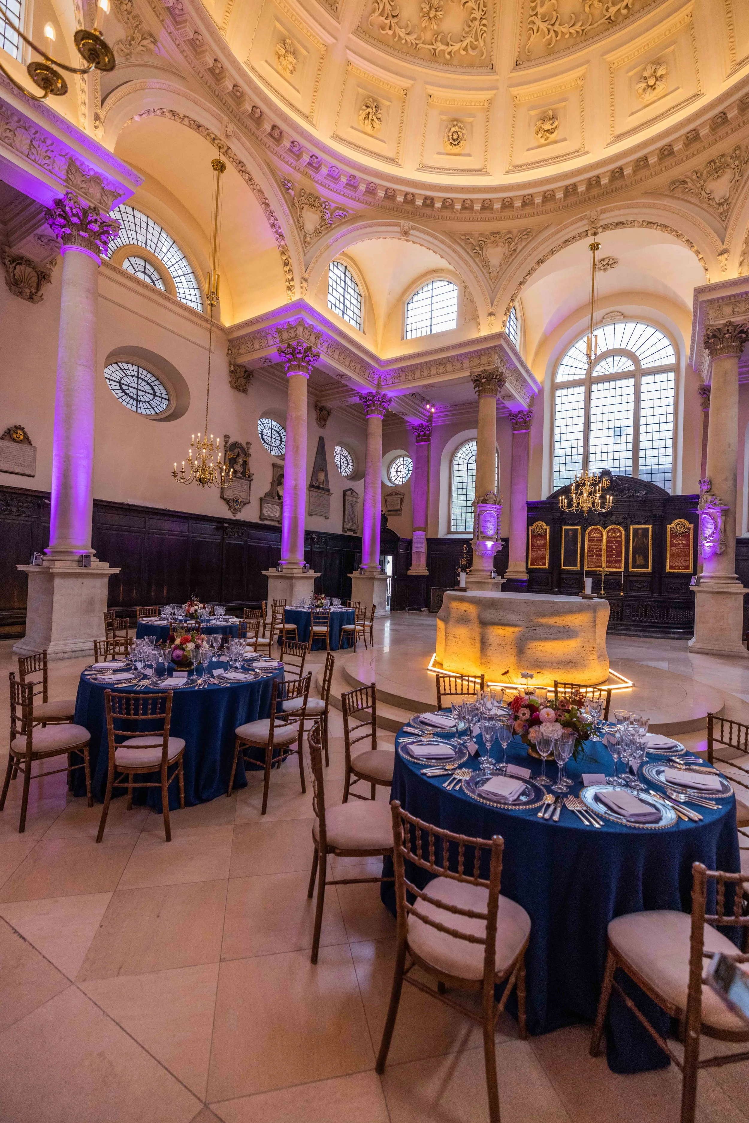 Elegant banquet hall with high arched ceilings, large stained glass windows, tall columns with purple lighting, chandeliers, and decorated tables with navy blue tablecloths and floral centerpieces.