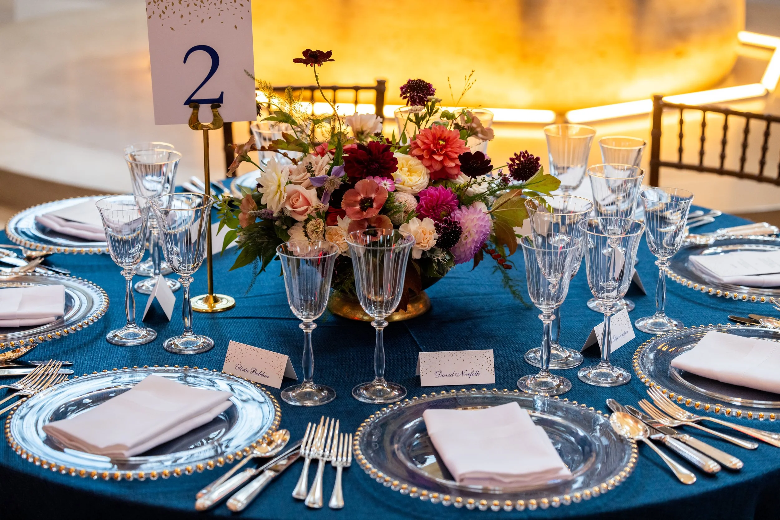 Elegant table setting with blue tablecloth, floral centerpiece, crystal glasses, silverware, and place cards at a banquet or wedding reception.