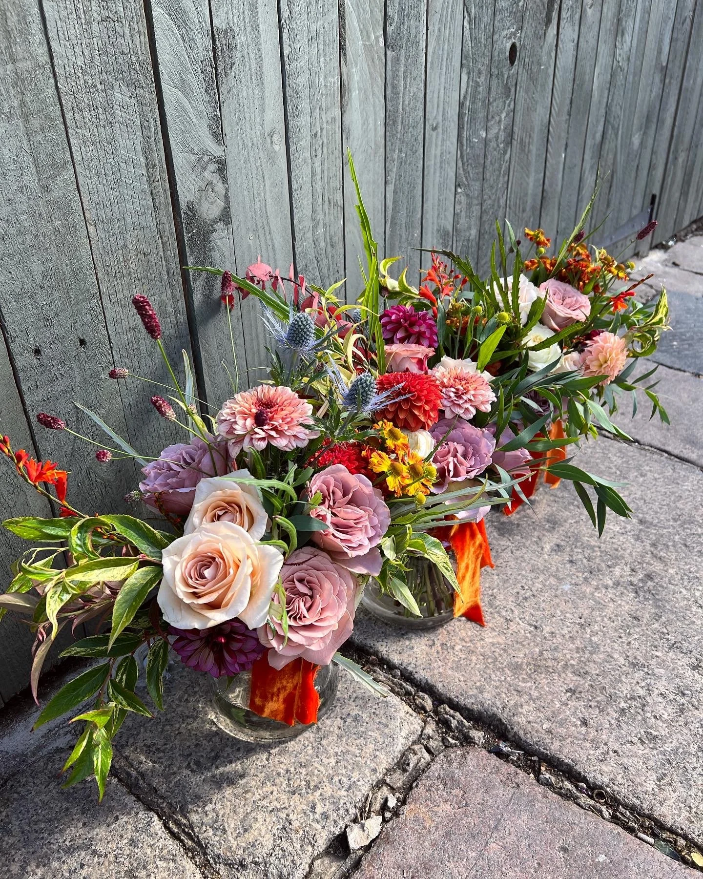 Two glass vases filled with assorted colorful flowers, including roses, dahlias, and other blooms, placed outdoors on a stone sidewalk against a wooden fence.