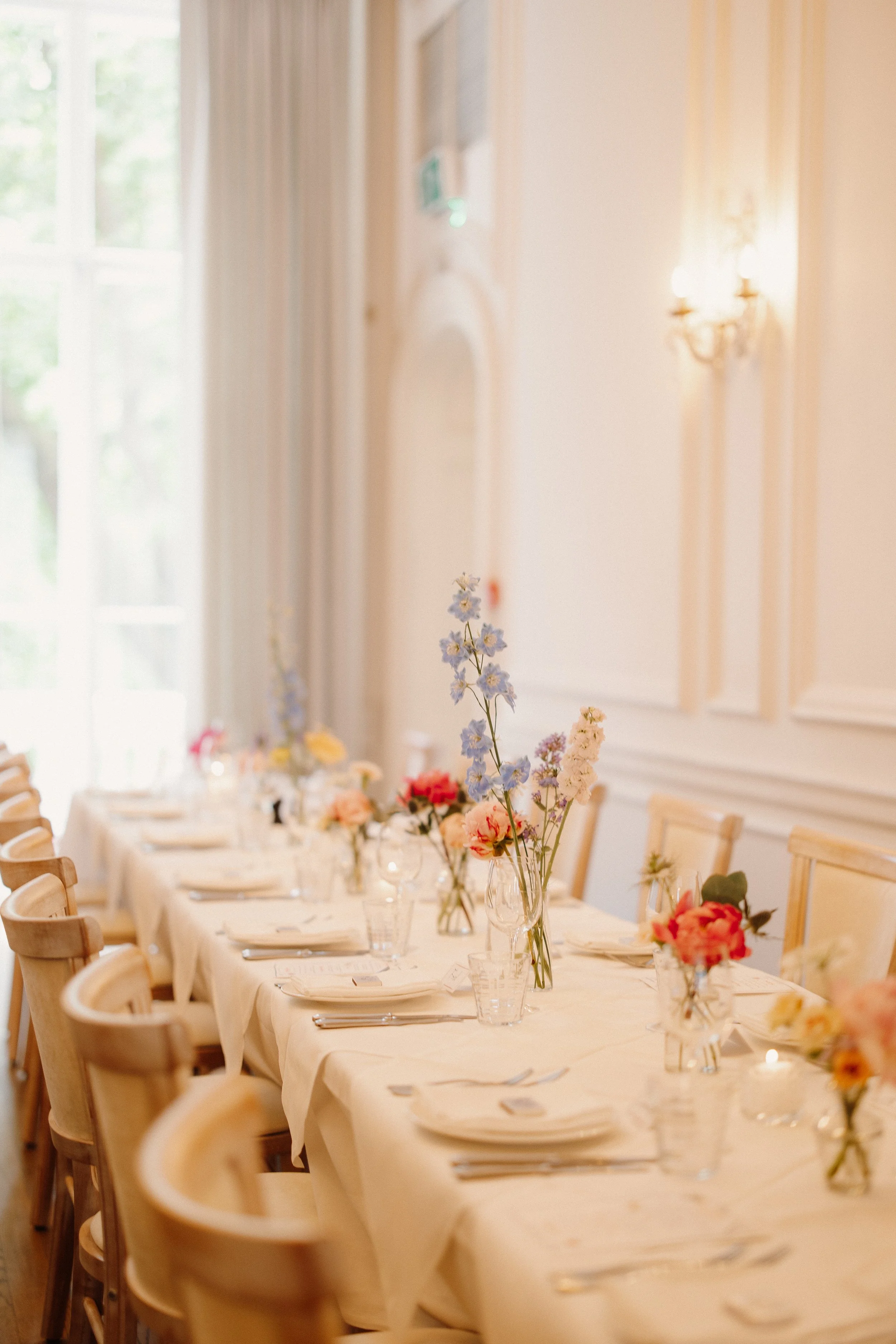 A long dining table set for a formal occasion with floral centerpieces and place settings, in a bright, elegant room with cream-colored walls and large windows.