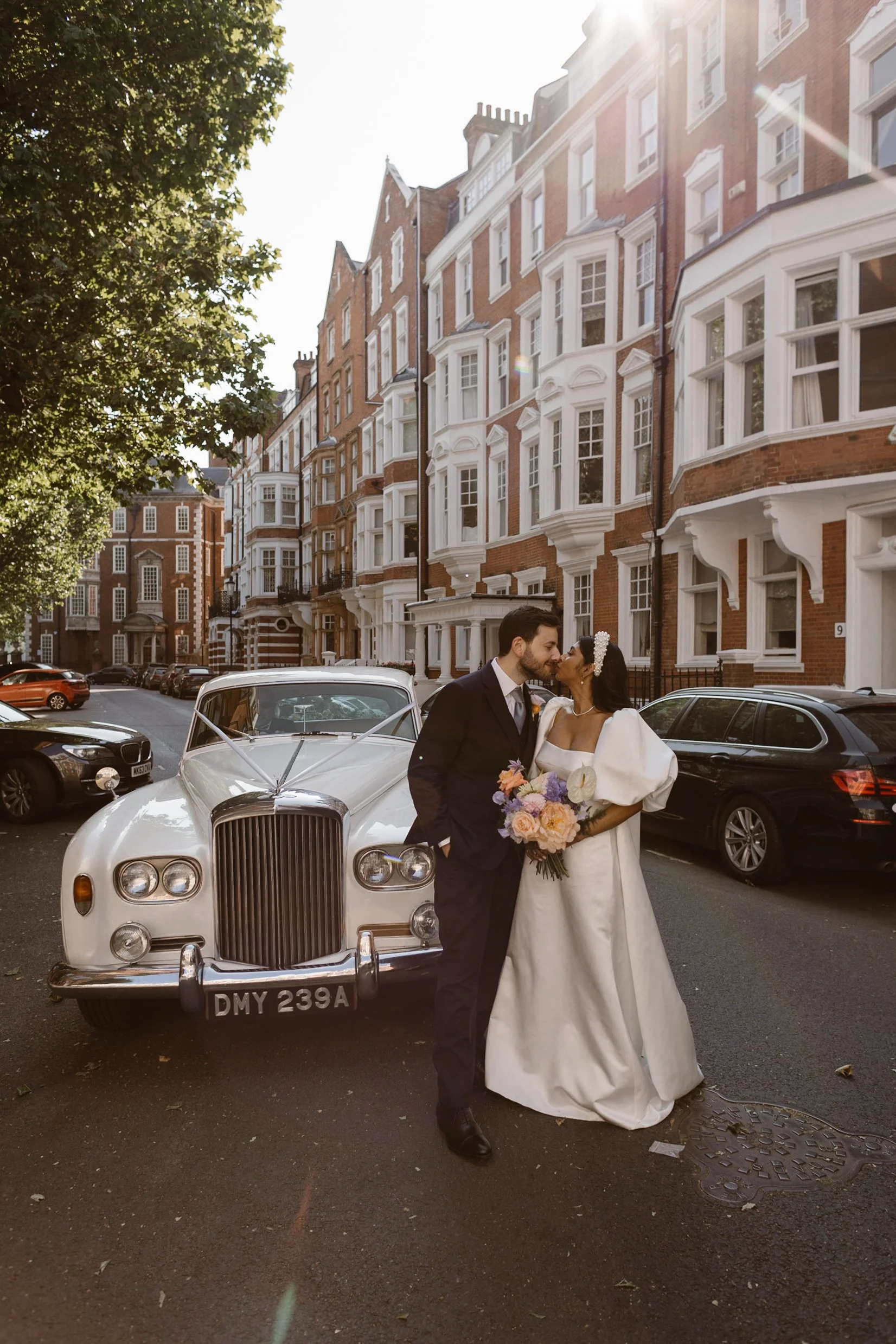 A newlywed couple sharing a kiss on a city street, standing in front of a vintage white car, with red brick Victorian-style buildings in the background, sunlight shining down.