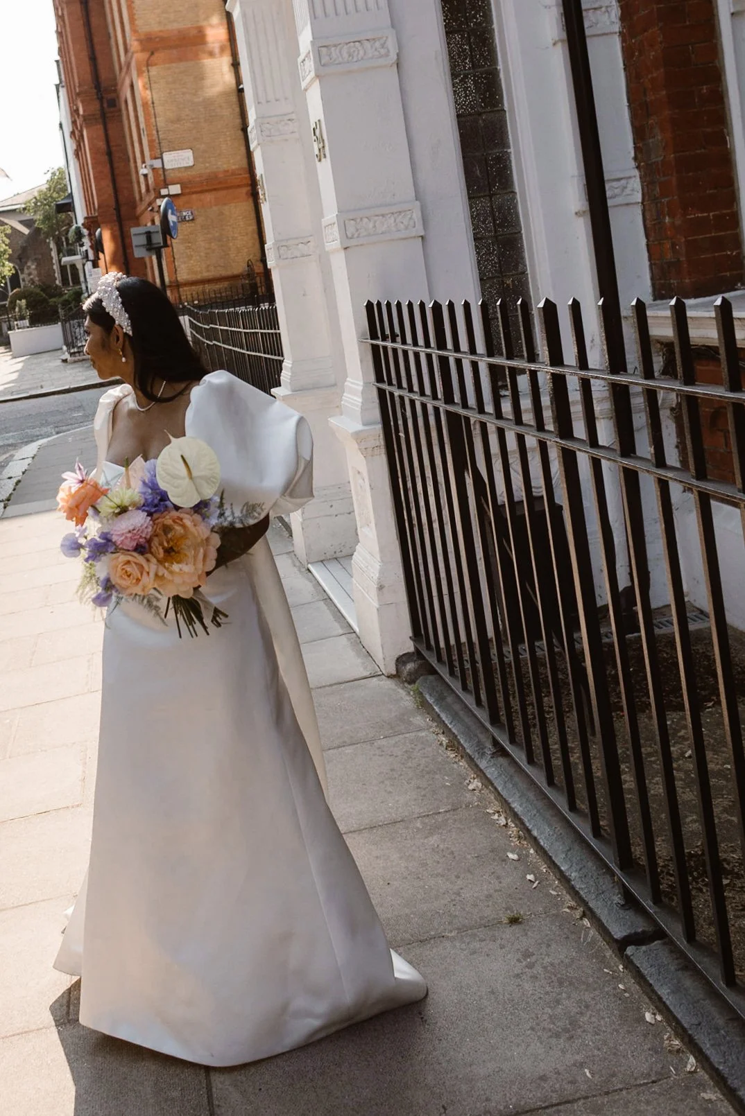 A woman in a white wedding gown holding a bouquet of flowers, standing on a city sidewalk.