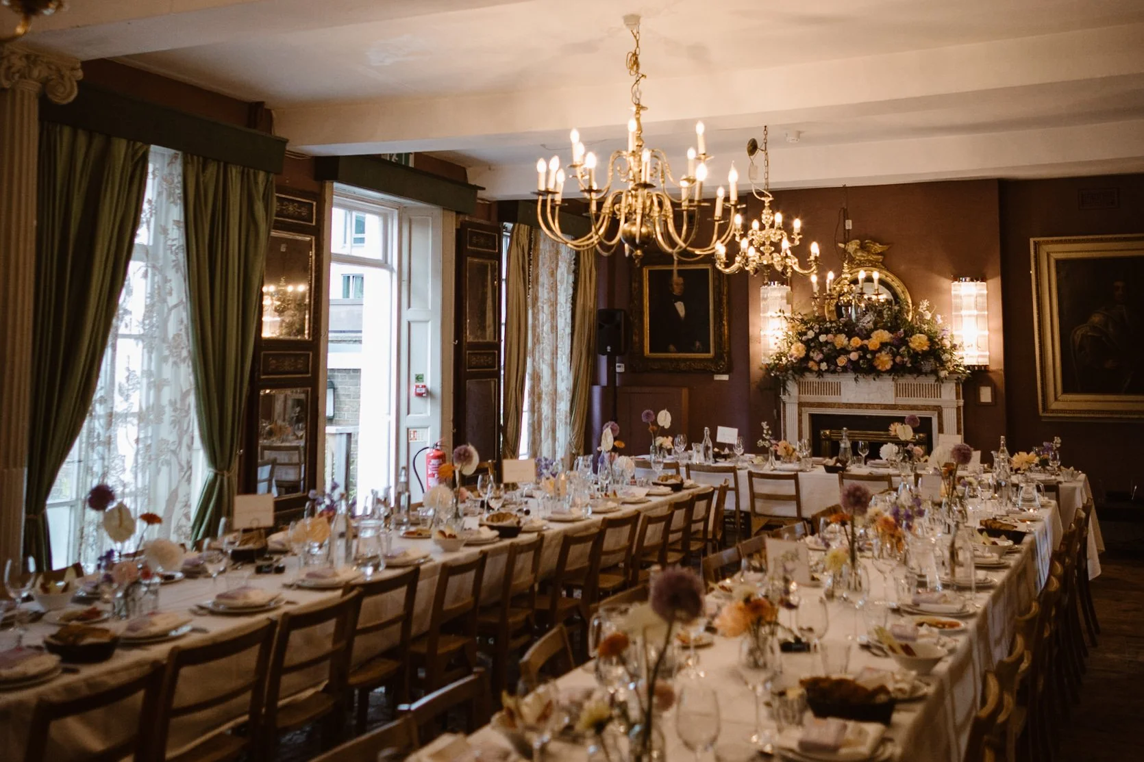 Elegant dining room set for a formal event, with long tables covered in white tablecloths, decorated with floral centerpieces, glassware, and place settings. The room has large windows with green curtains, ornate chandeliers hanging from the ceiling,