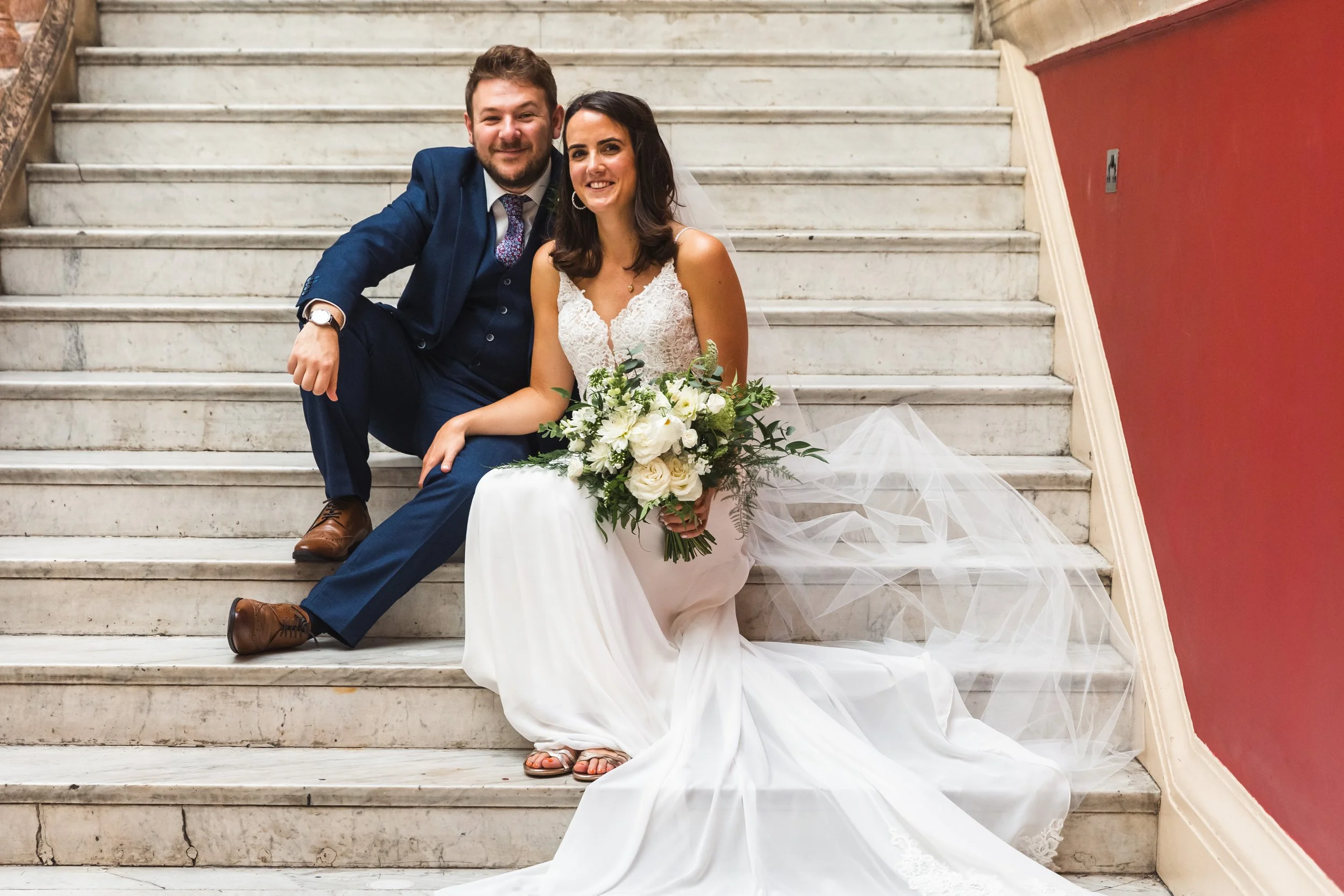 A bride and groom sitting on a staircase during their wedding, with the bride holding a bouquet of white flowers and greenery, and the groom dressed in a blue suit.