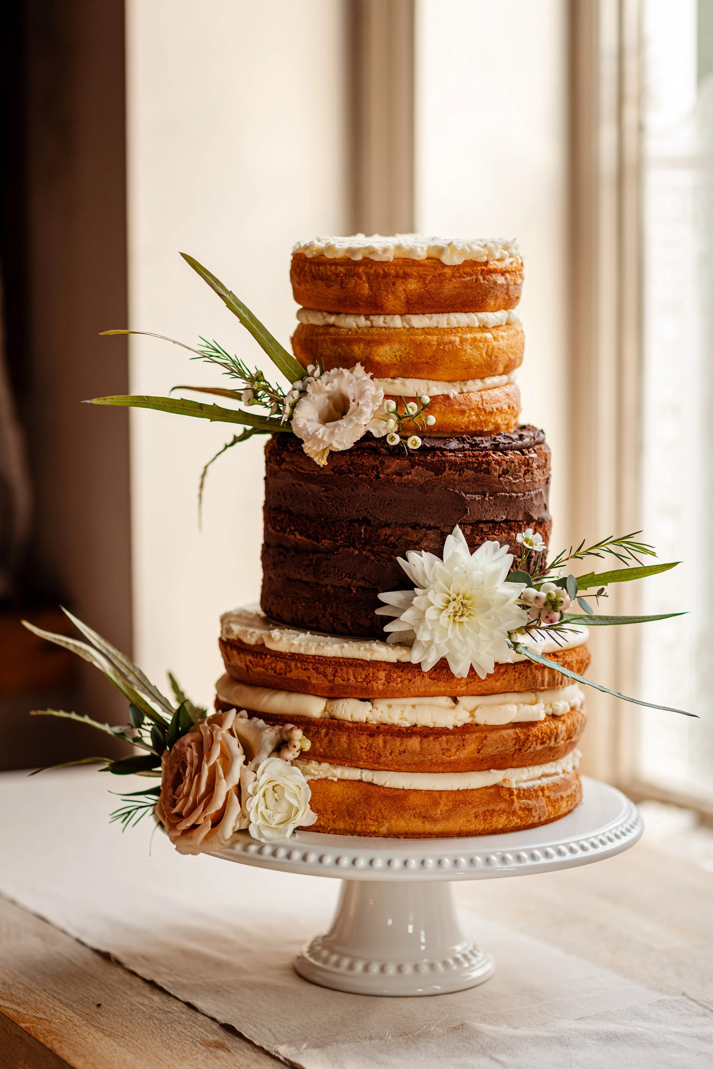 A three-tier naked wedding cake decorated with white, beige, and pink flowers and greenery, sitting on a white cake stand in front of a window.