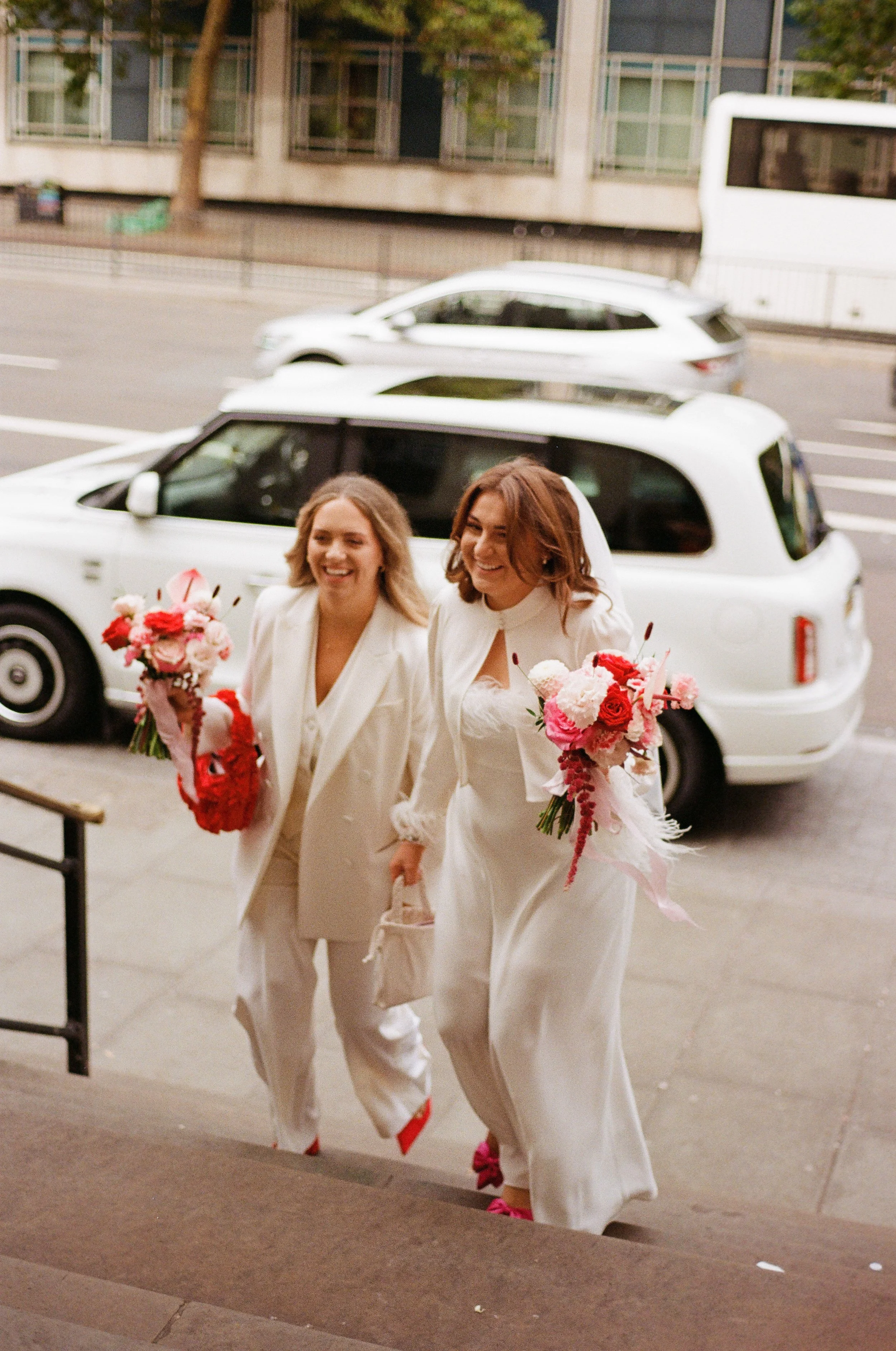 Two women in white wedding attire, holding bouquets, walking up stairs outside a building, smiling and happy.