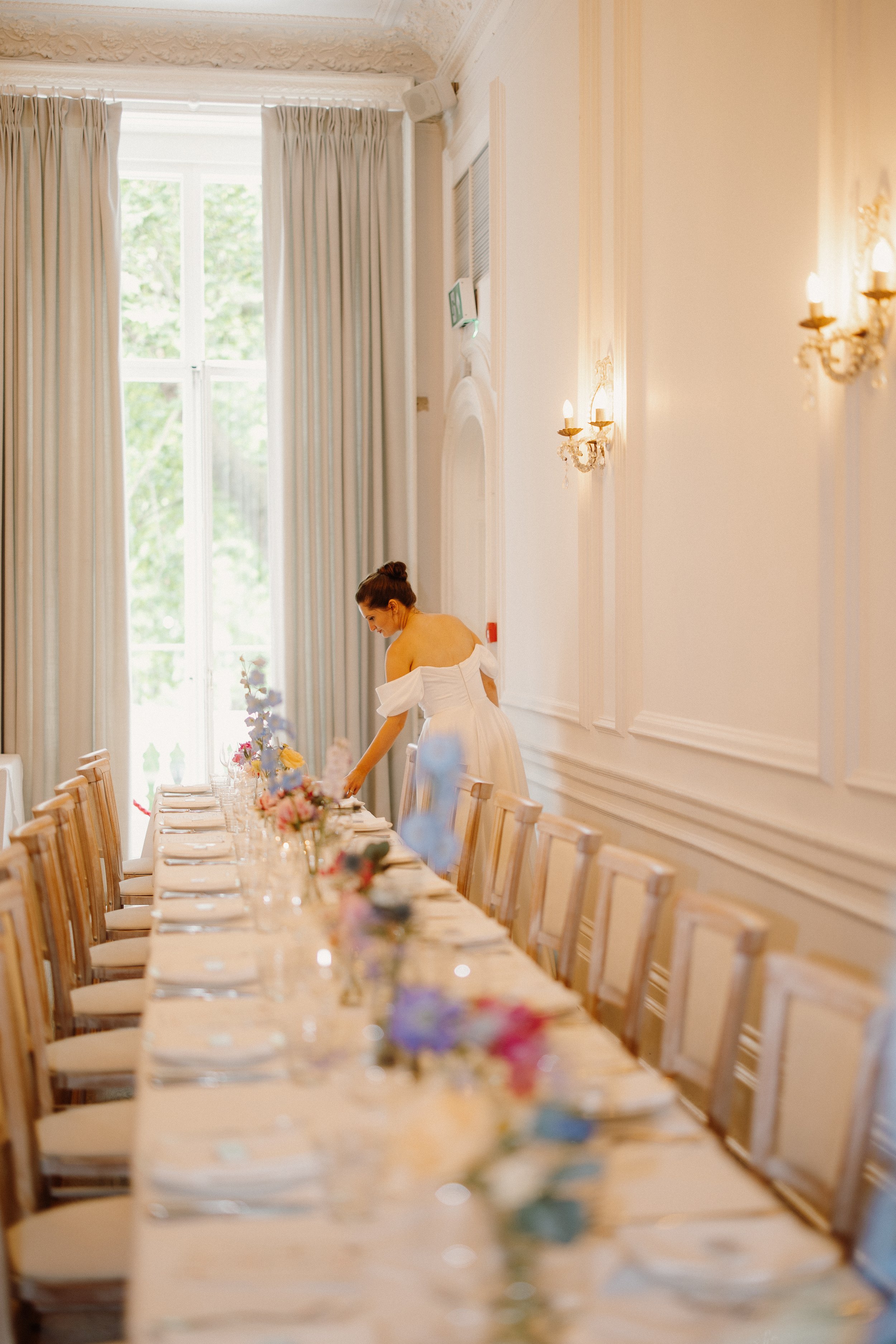 A woman in a white dress arranging flowers on a long banquet table in a bright, elegant room with large windows and cream-colored walls.