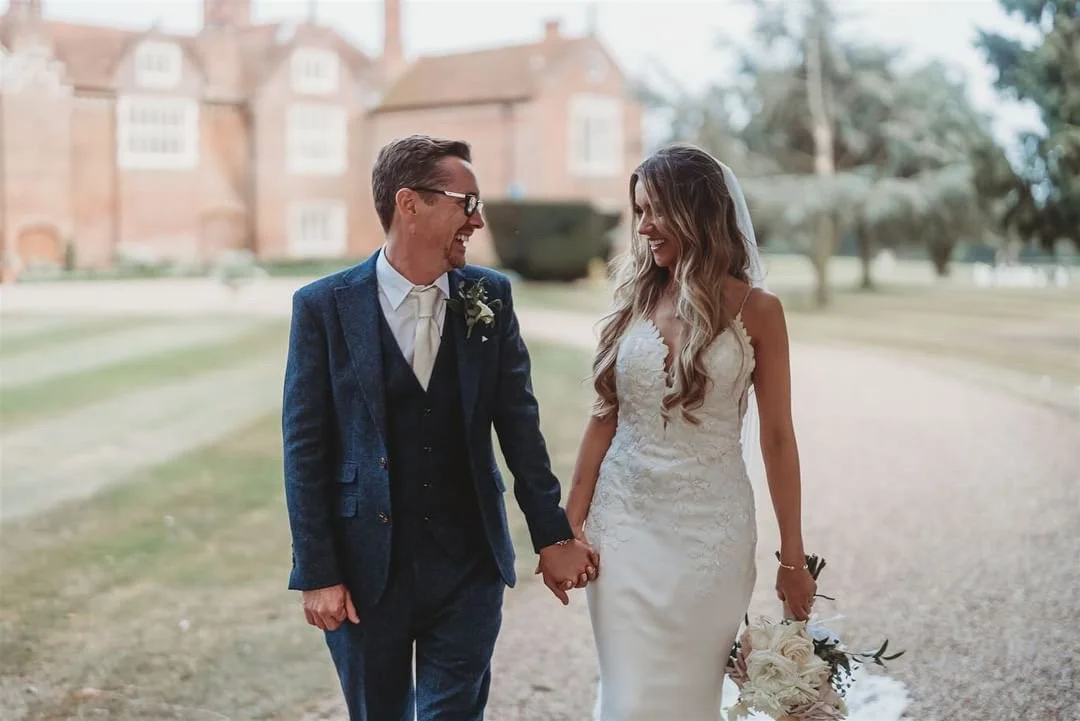 A bride and groom holding hands and smiling at each other outdoors on a wedding day.