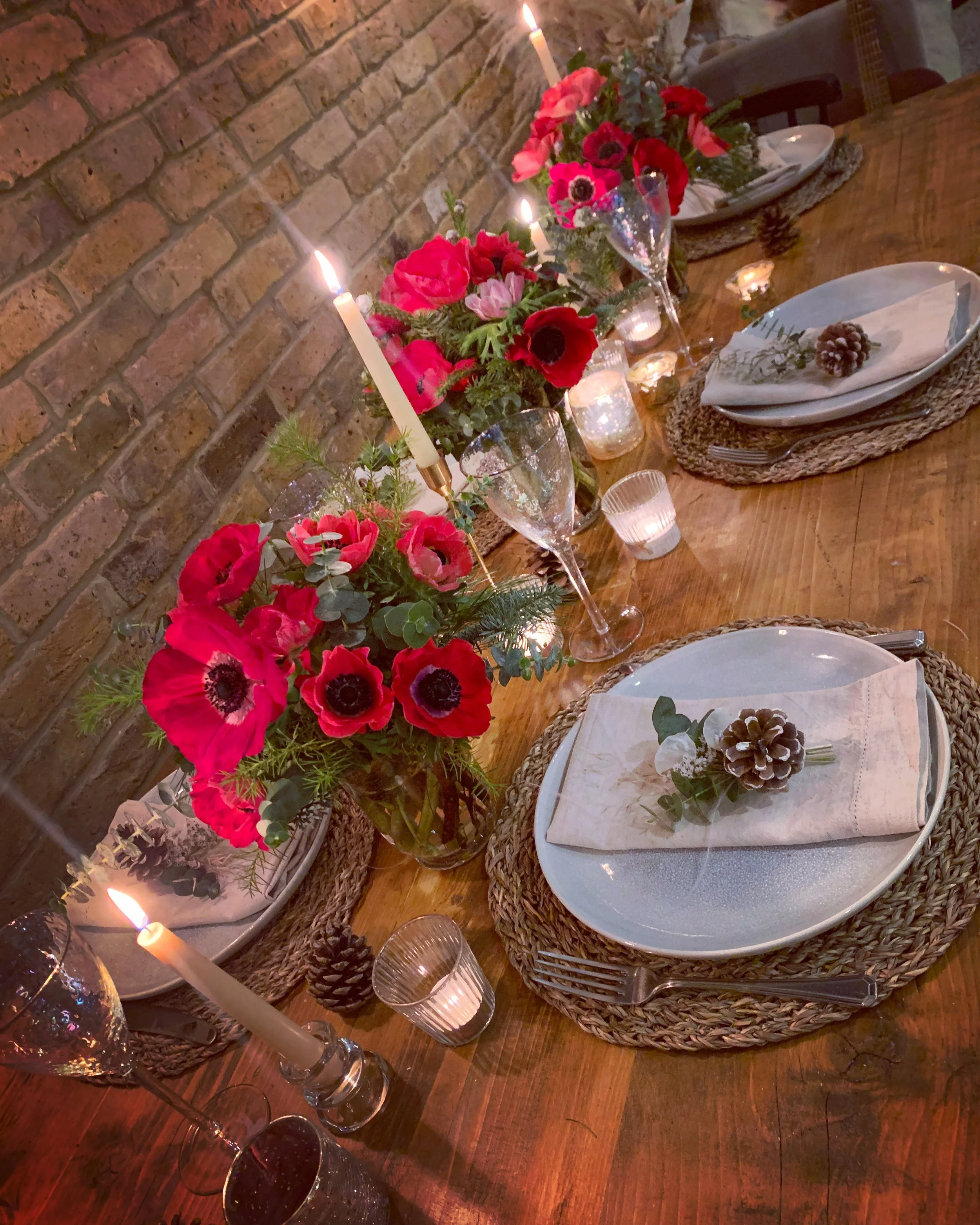 A decorated dinner table with pink and red flowers, lit candles, tableware, and pine cone decorations.