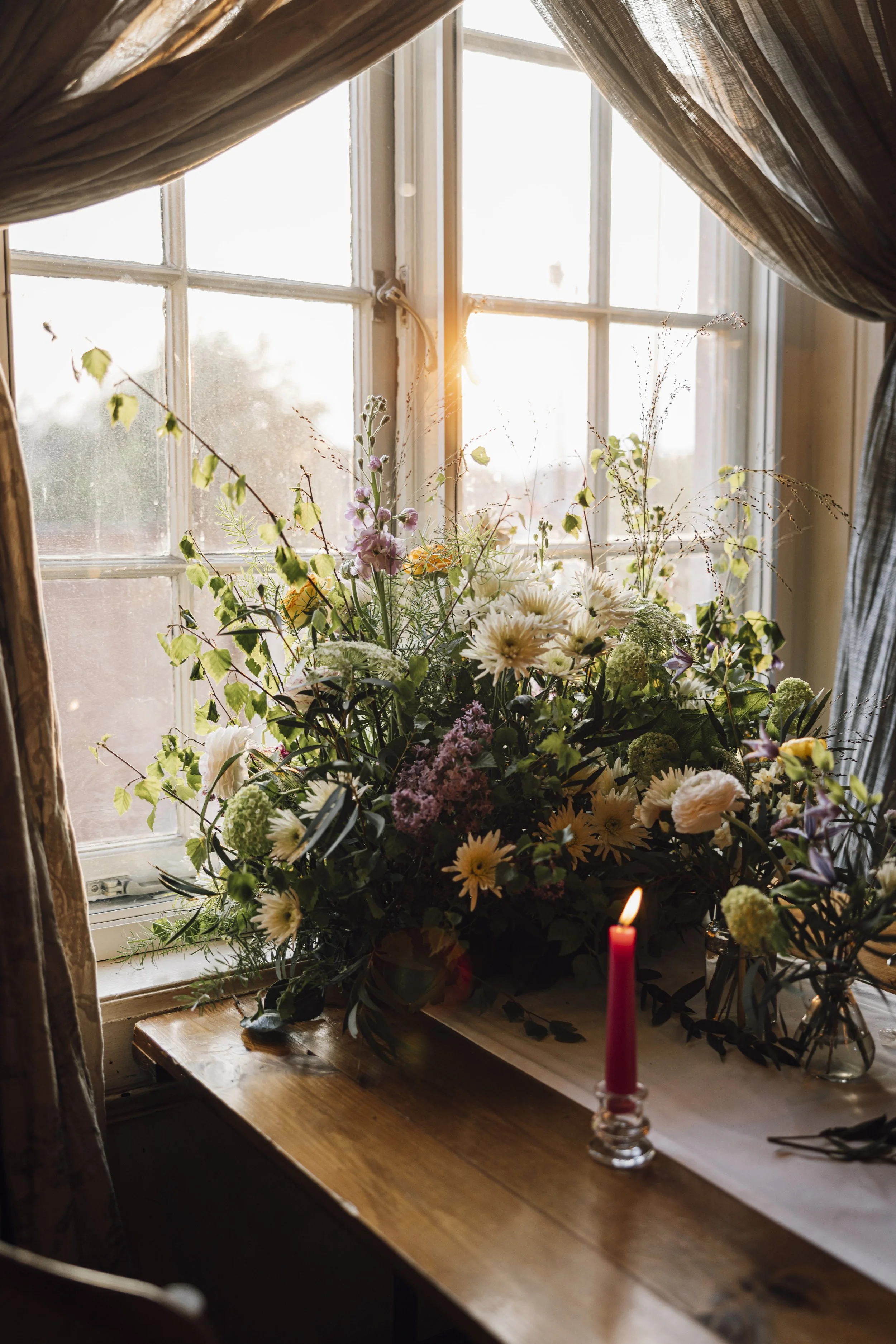 A window sill with a large floral arrangement illuminated by sunlight, and a single lit pink candle.