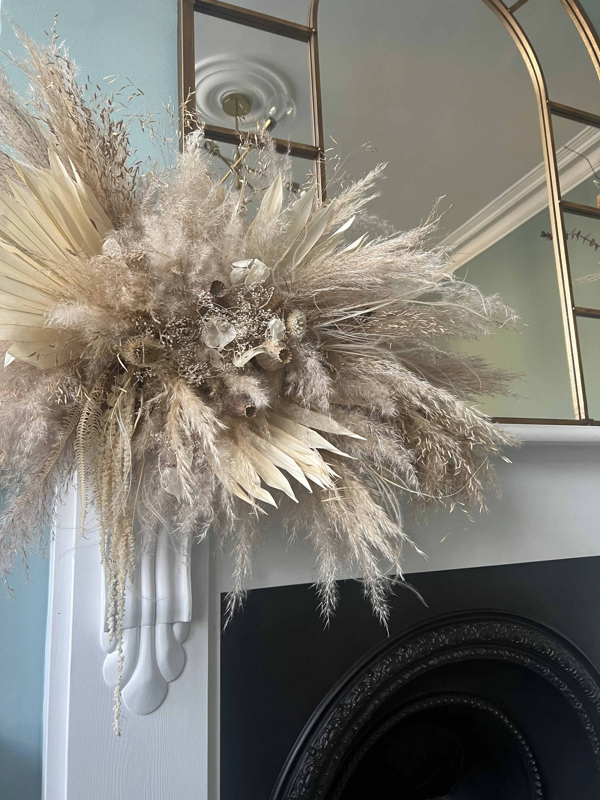 Dried floral arrangement above a fireplace mantel, featuring pampas grass, palm leaves, and other dried botanicals in neutral tones.