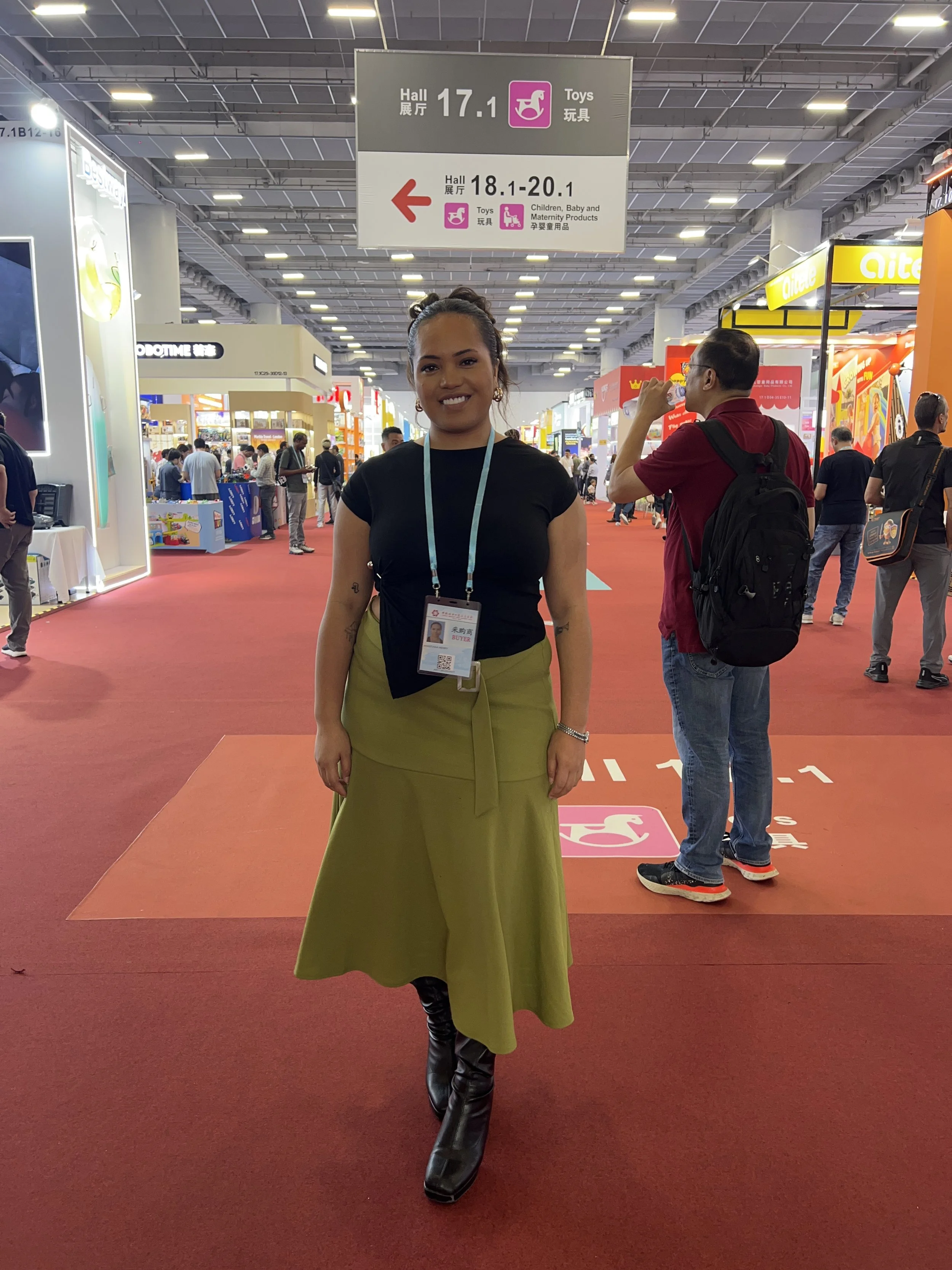 A woman standing on a red carpet inside a convention center, smiling at the camera. She is wearing a black top, an olive green skirt, and black boots. Around her neck is a blue lanyard with an ID badge. In the background are other people walking and 