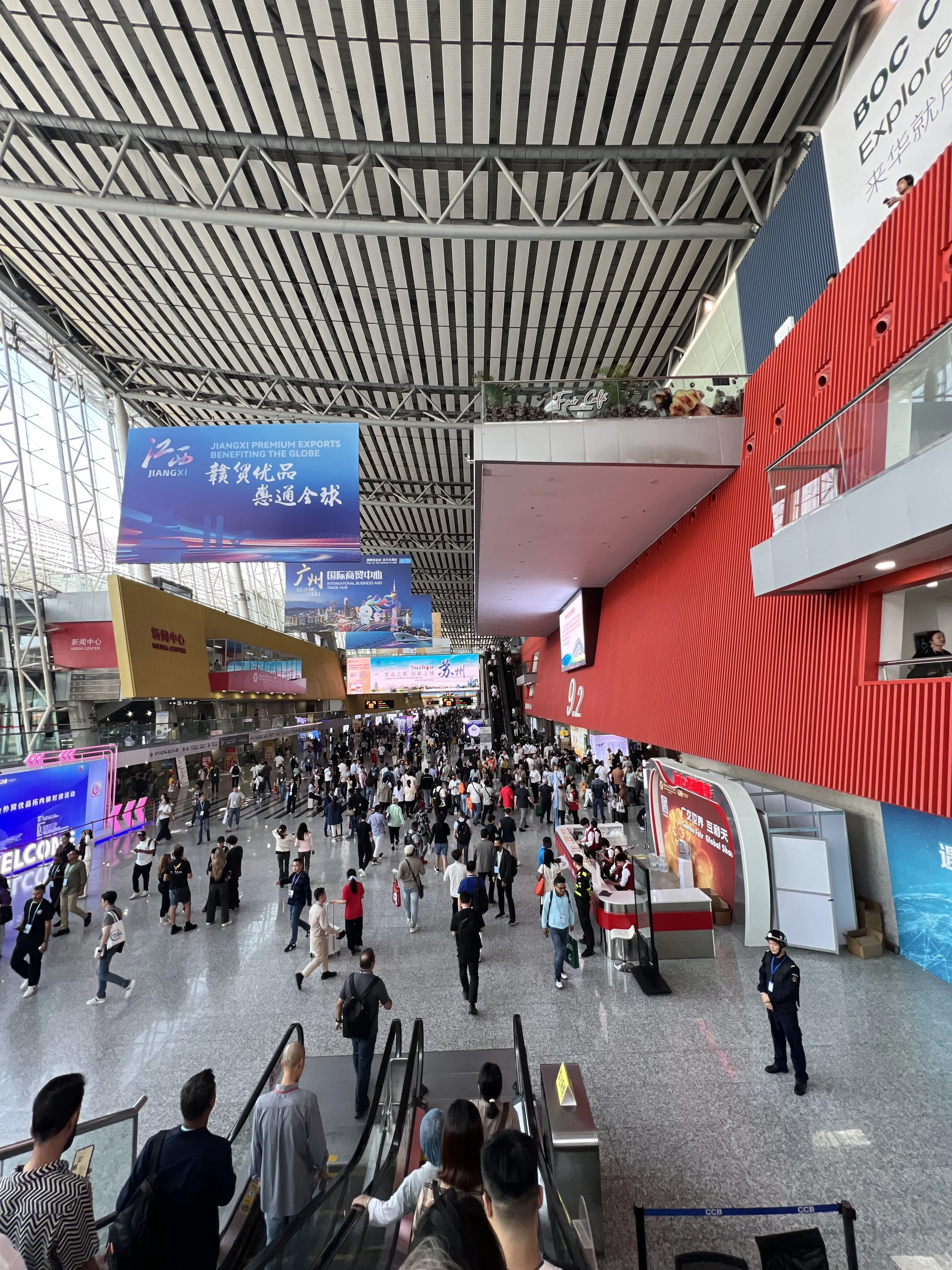 Crowded airport terminal with travelers walking and standing, escalator in foreground, large digital screens and advertisements on walls, high ceiling with structural beams, and a security personnel standing near the red wall.