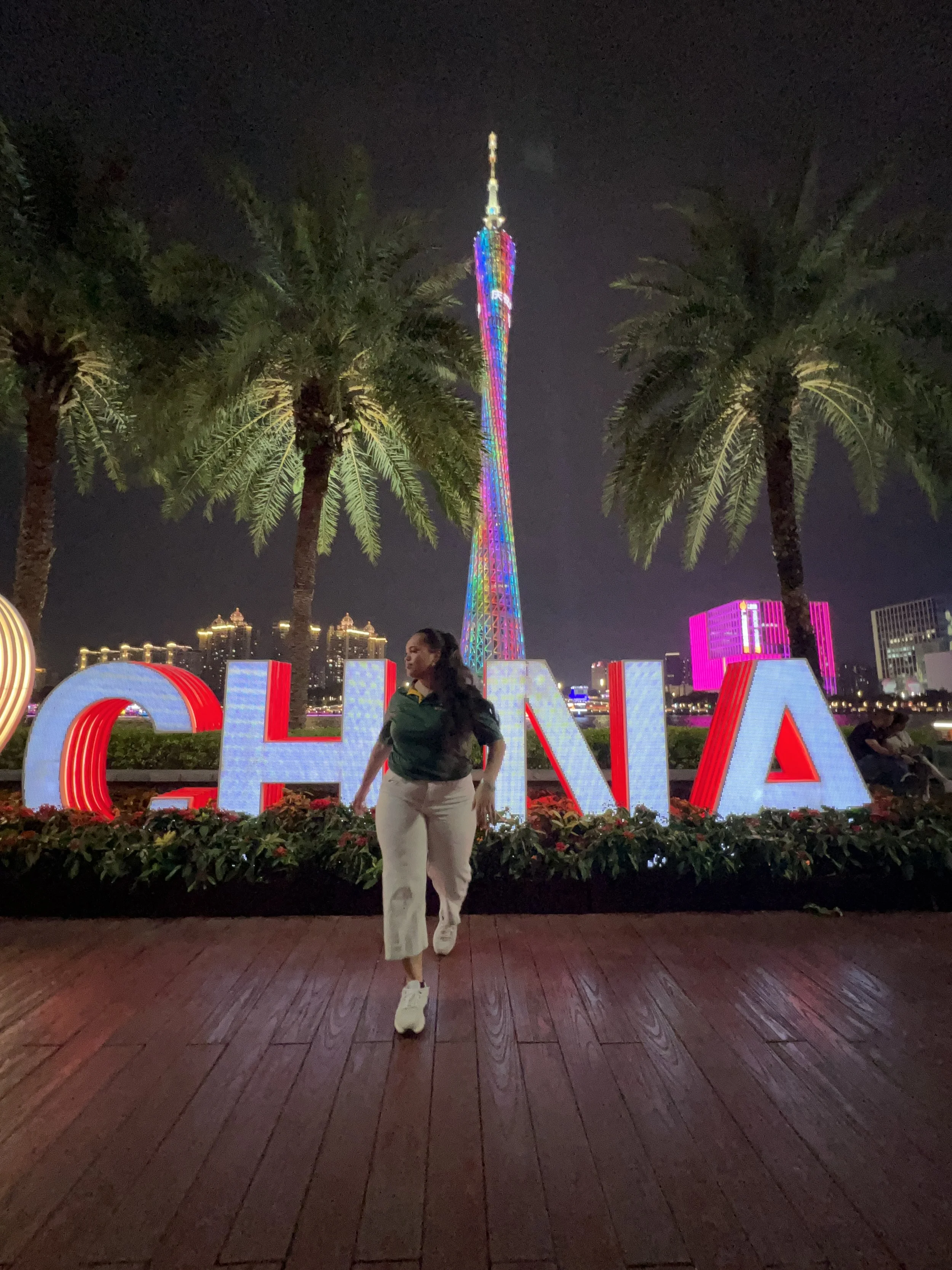 A woman walking on a wooden pathway at night in front of large illuminated letters spelling 'CHINA' with colorful neon lights, palm trees, and a tall, brightly lit tower in the background.