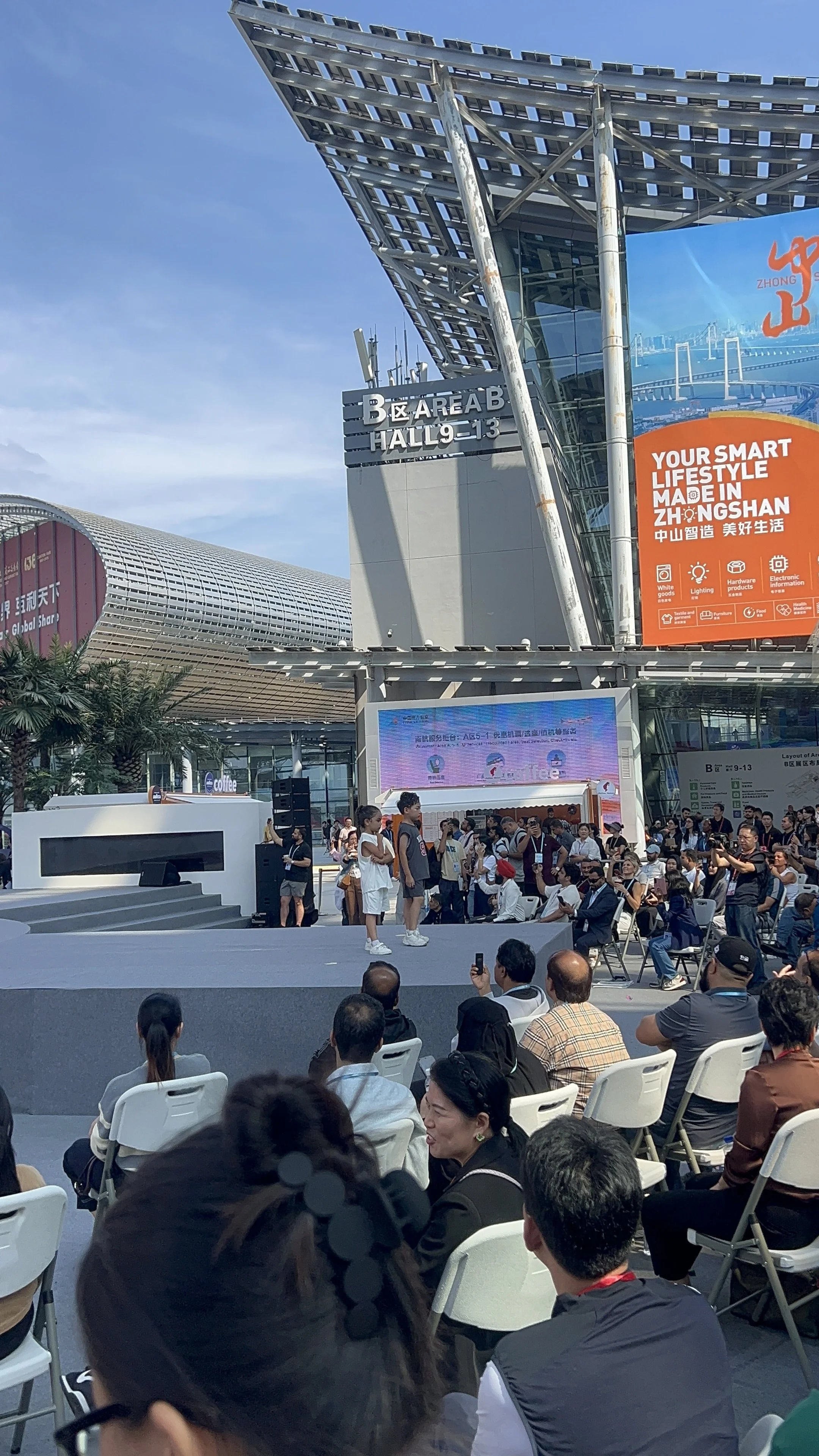 A crowd of people attending a performance on an outdoor stage at a convention center. The stage has children performing, with some individuals taking photos or videos. The background shows the modern architecture of the exhibition hall and a large di