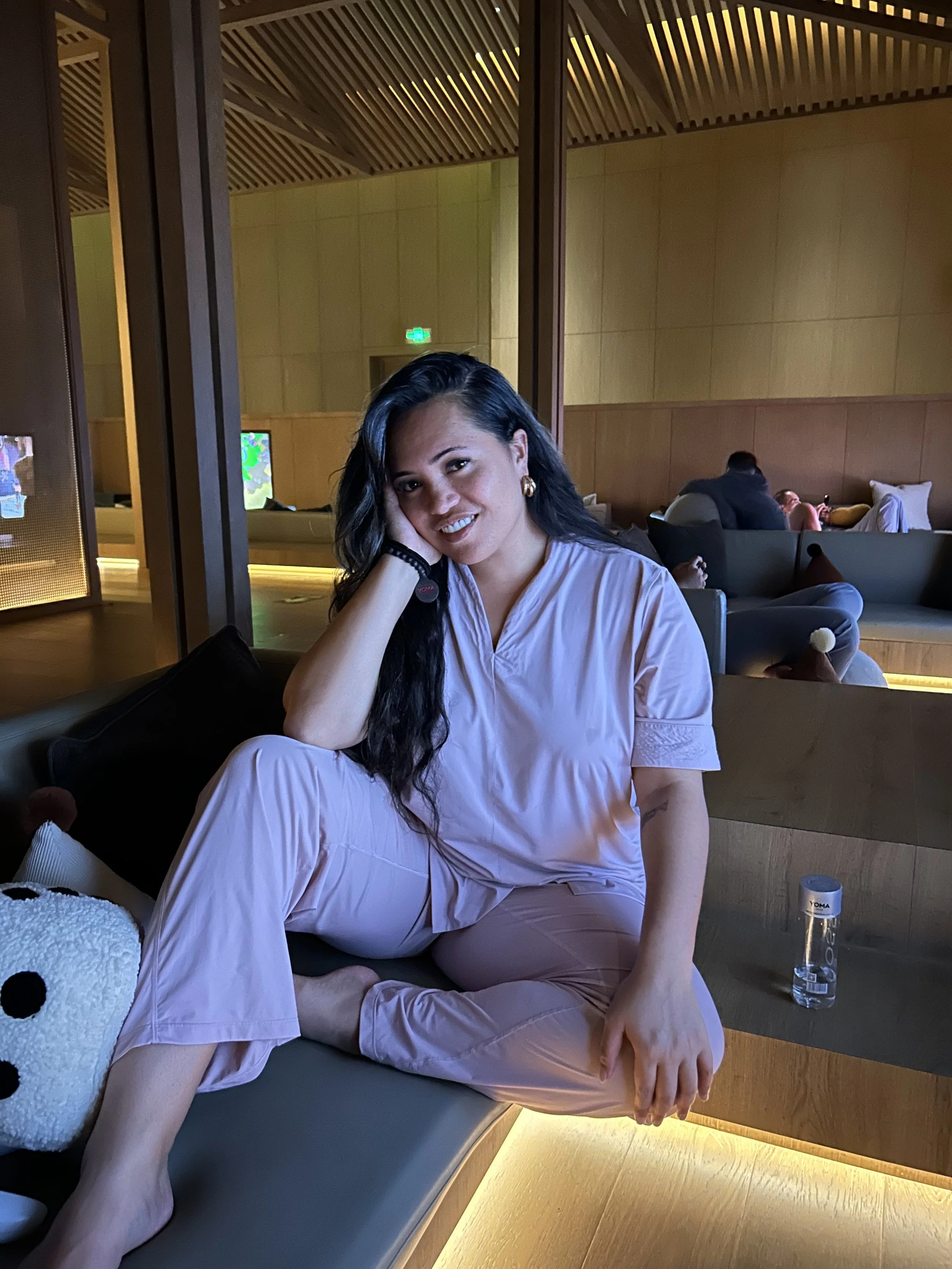 A woman with long dark hair in pink pajamas sitting on a black sofa, smiling and resting her head on her hand, in a warmly lit modern lounge.