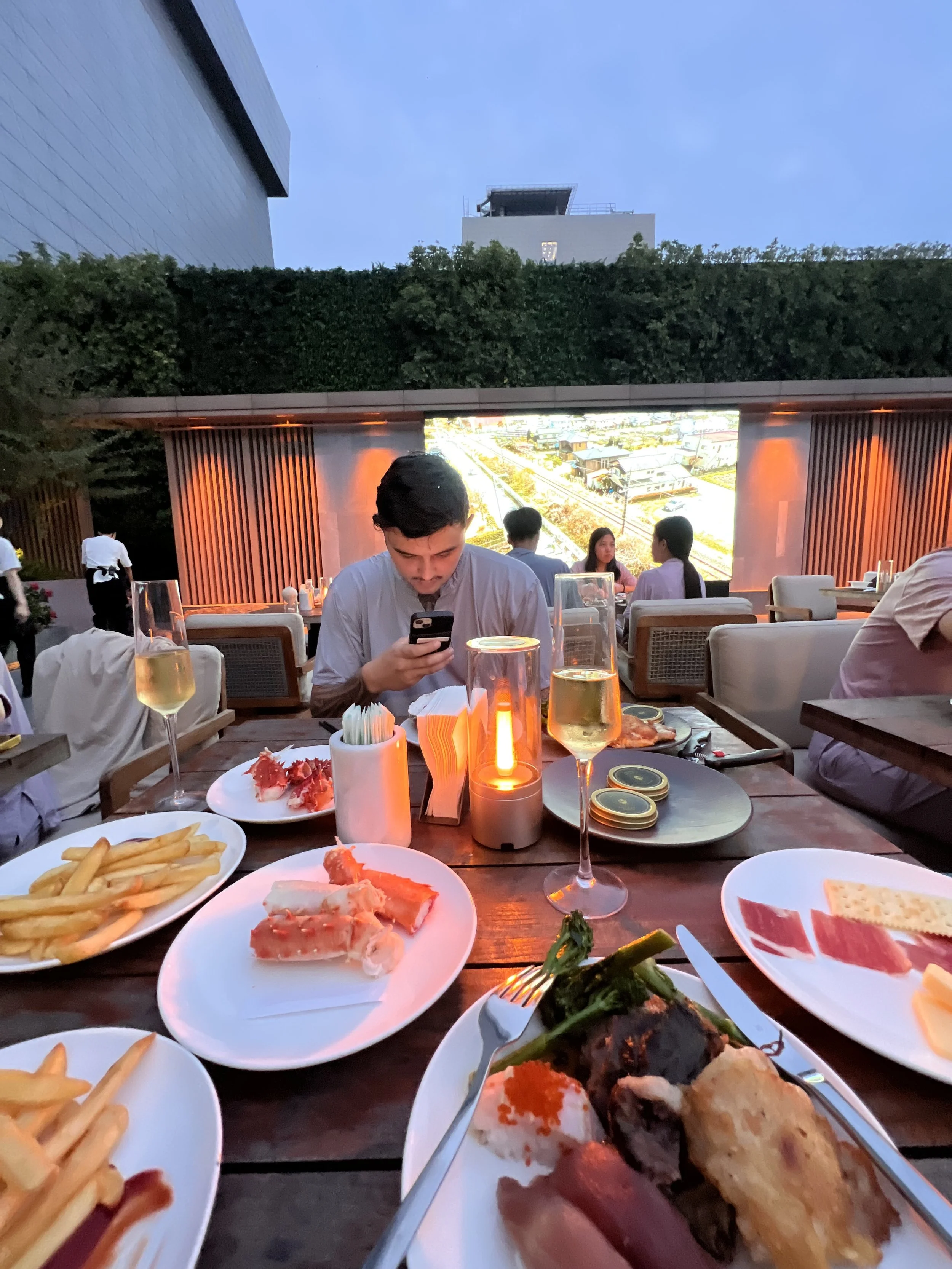 People dining at outdoor restaurant in evening with food and drinks on table, large screen displaying cityscape in background.