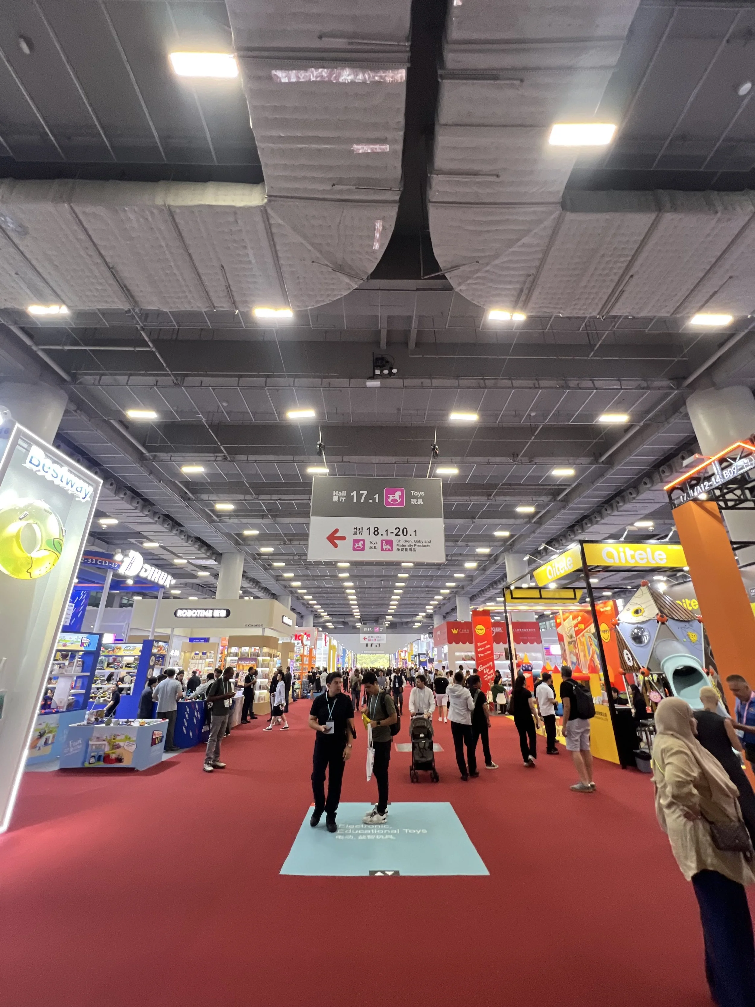 An indoor trade show or exhibition with multiple booths and visitors walking through. Signs hang from the ceiling indicating hall numbers and sections for toys and children’s products.