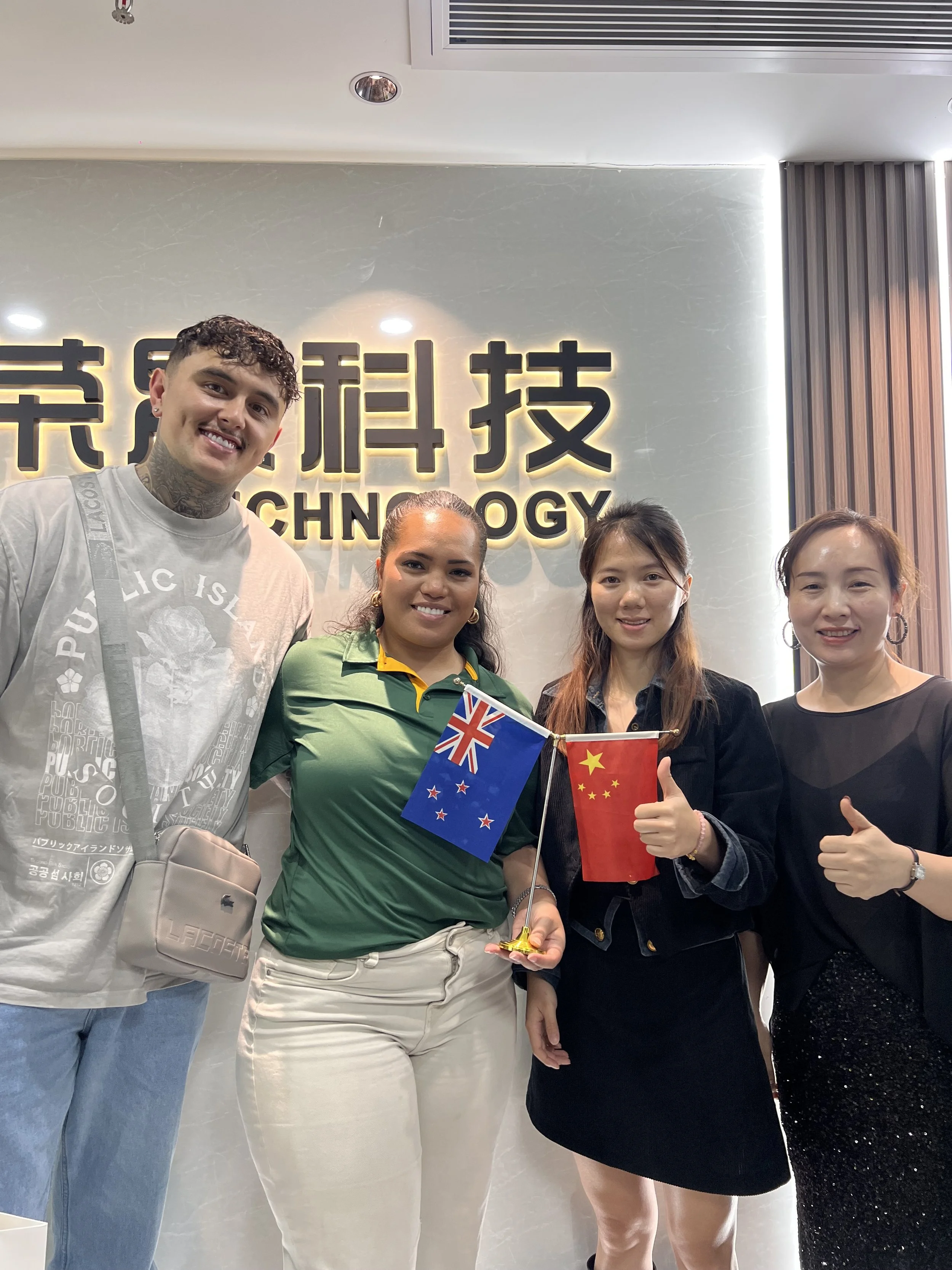 Four people standing together in front of a wall with the word 'Technology' in large letters. The person second from the left is holding New Zealand and Chinese flags, and the person second from the right is giving a thumbs-up sign.