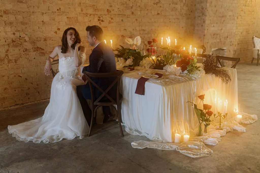 Bride wearing an off-the-shoulder satin wedding gown seated beside a candlelit reception table with red roses and romantic styling at The Loft on Hindley in Adelaide.