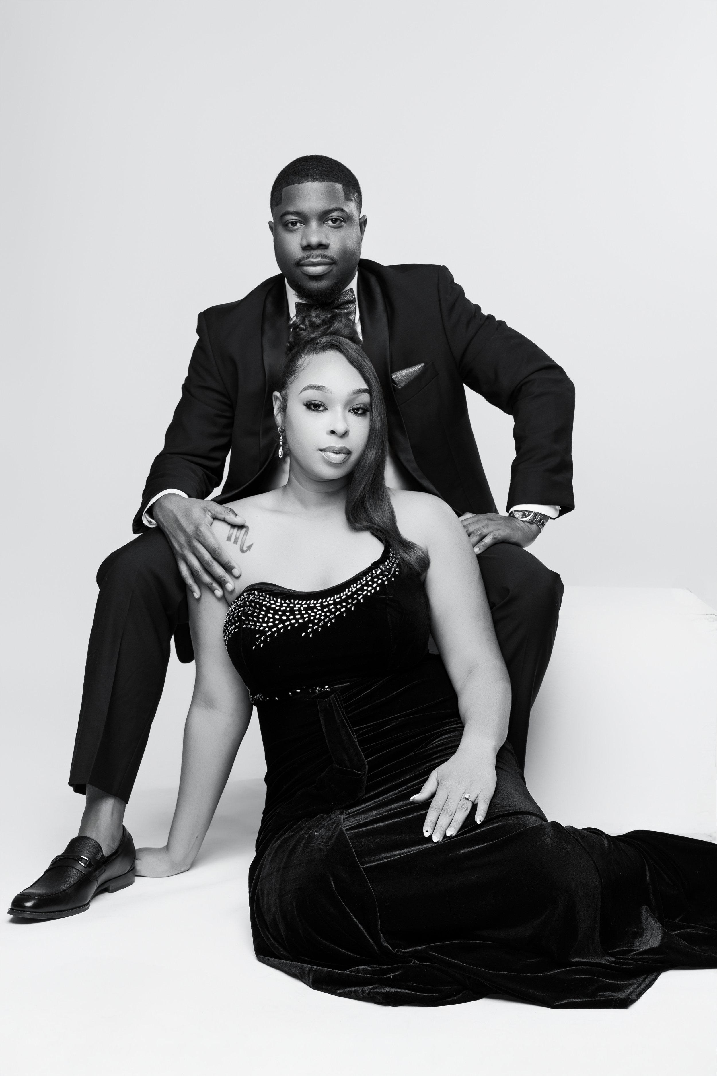 Black and white photo of a man in a tuxedo standing behind a woman in a strapless evening gown, seated on a white surface, both looking at the camera.