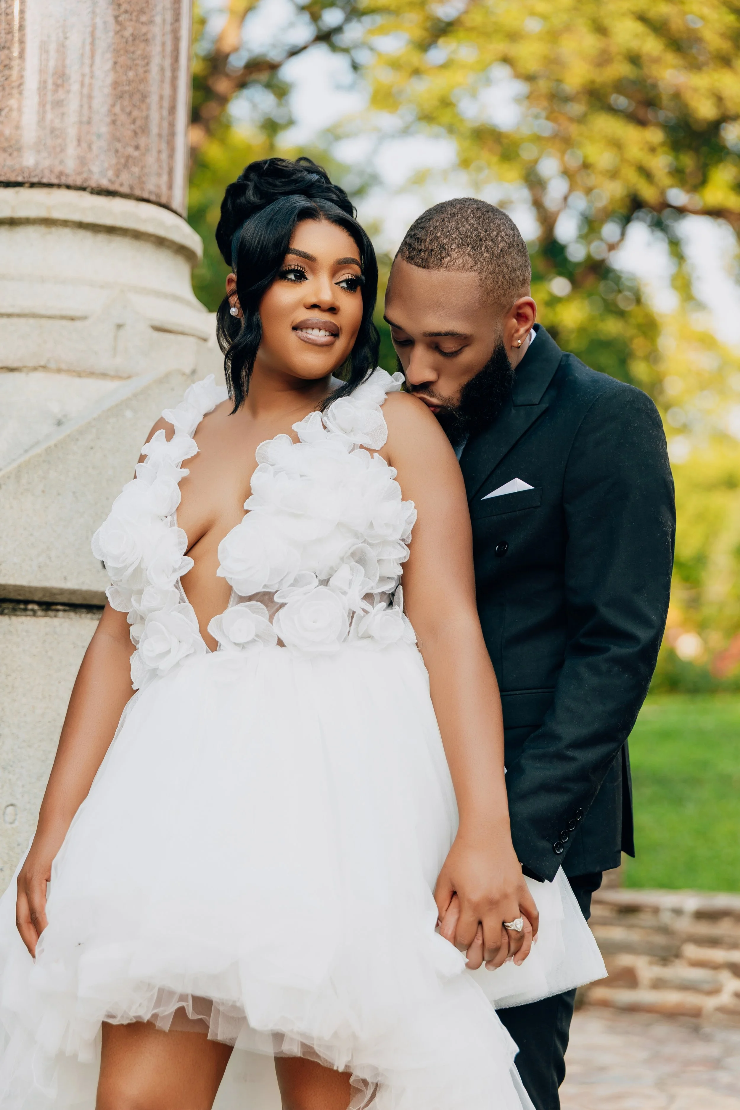 A bride in a white dress with floral details stands next to a groom in a black suit in a park setting.