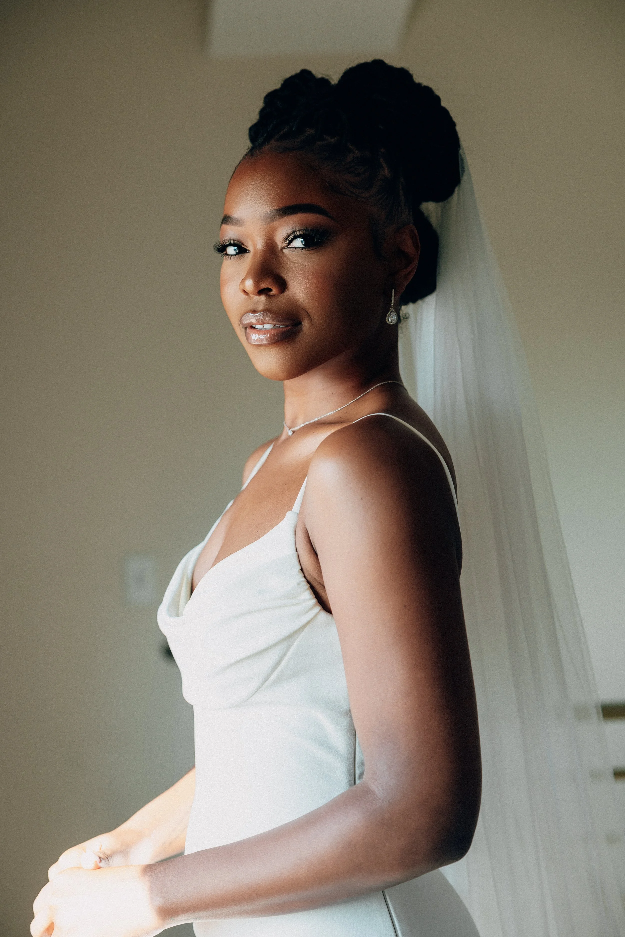 Bride in a white wedding gown with a veil, looking at the camera, indoors with plain background.