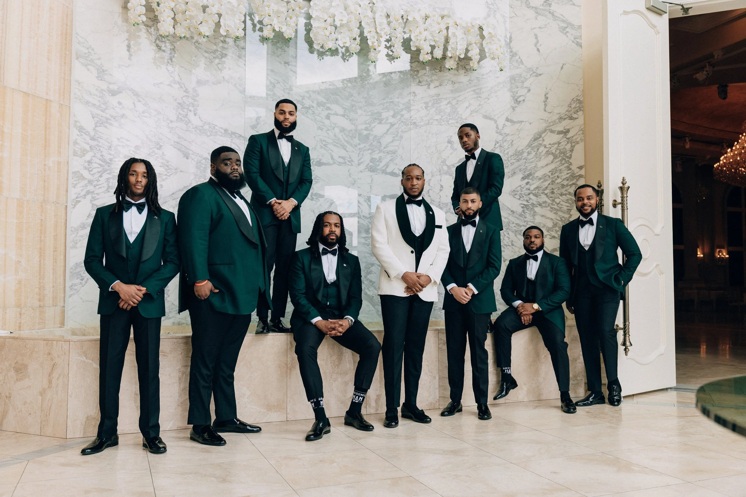 A group of ten men dressed in tuxedos, some with bow ties and some with black shirts, gathered in an elegant indoor setting with a marble wall and white floral arrangements in the background.
