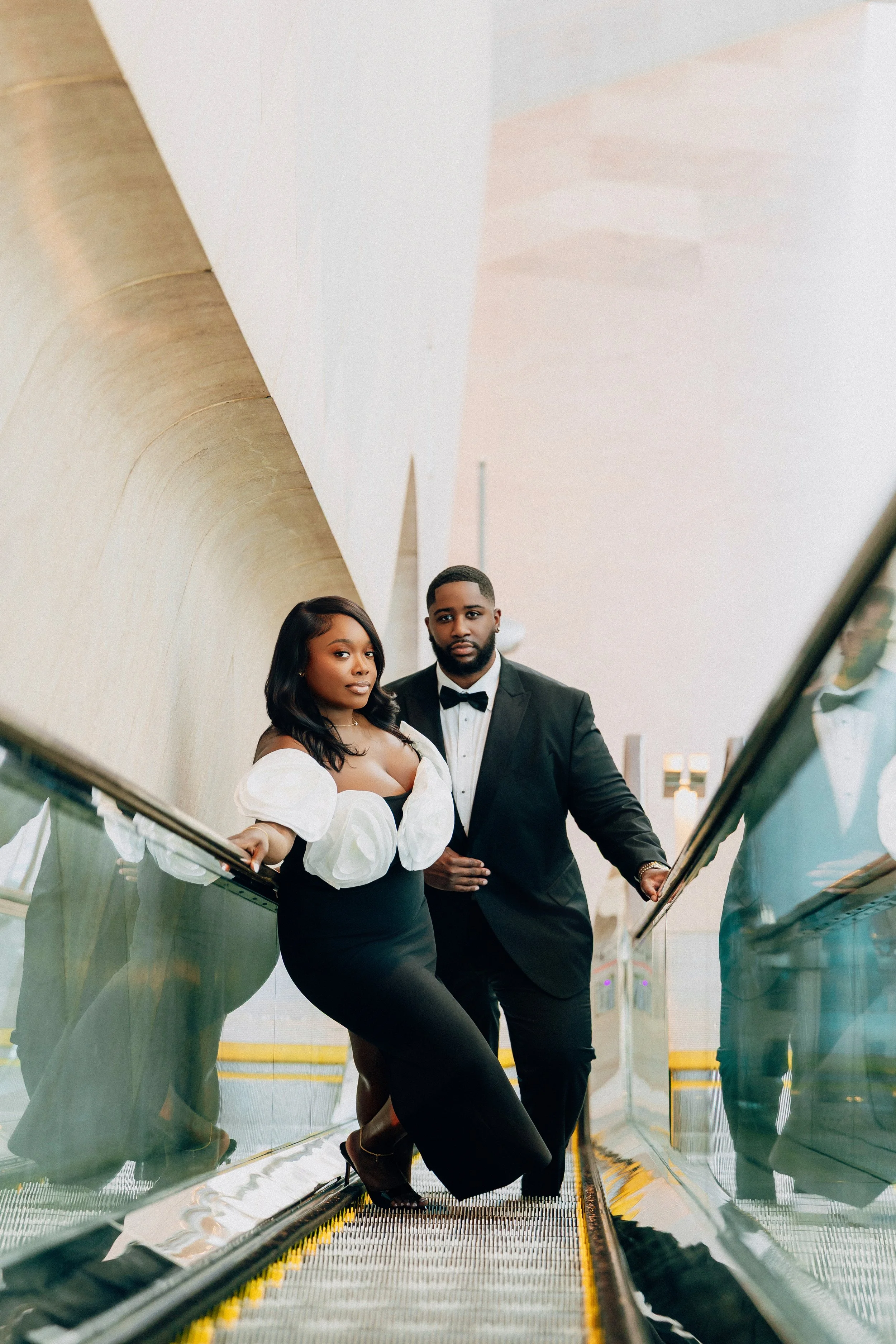 A couple dressed in formal attire, standing on an escalator. The woman wears a black dress with puffy white sleeves and black heels, while the man wears a black tuxedo with a bow tie.